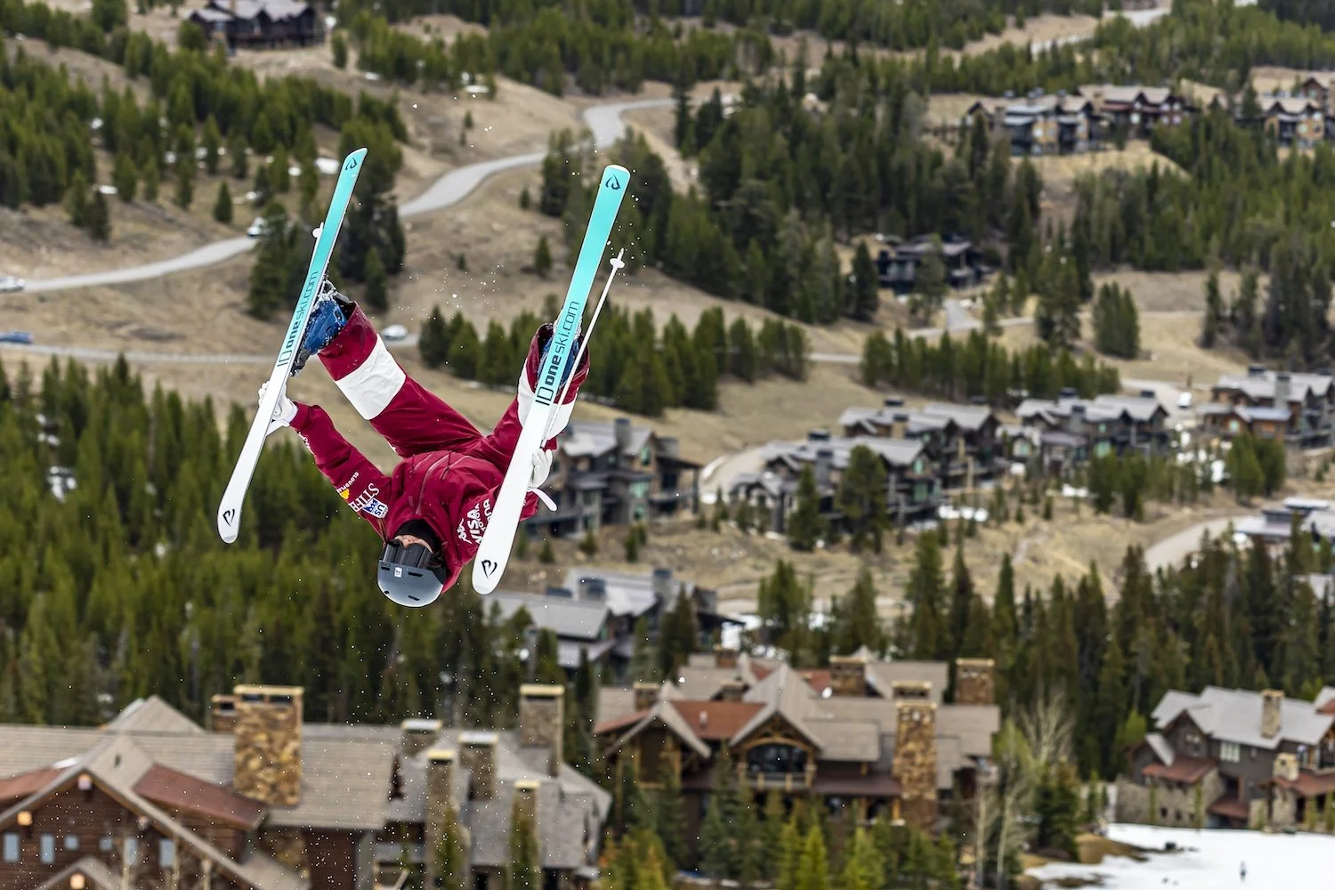 A skier executing a mid-air stunt above a mountain town, dressed in red ski gear, with a background of trees and houses.