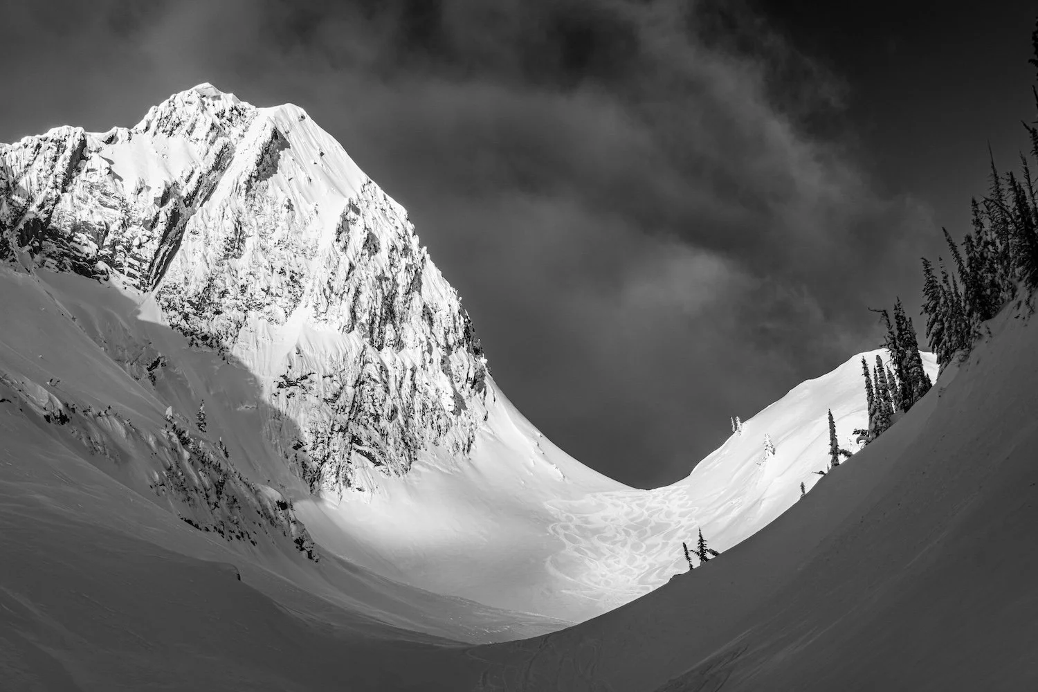 Black and white photograph of a snow-covered mountain range, with a steep peak on the left and a valley with snow tracks in the foreground, and pine trees on the right side. Island Lake Lodge. Fernie, British Columbia, Canada