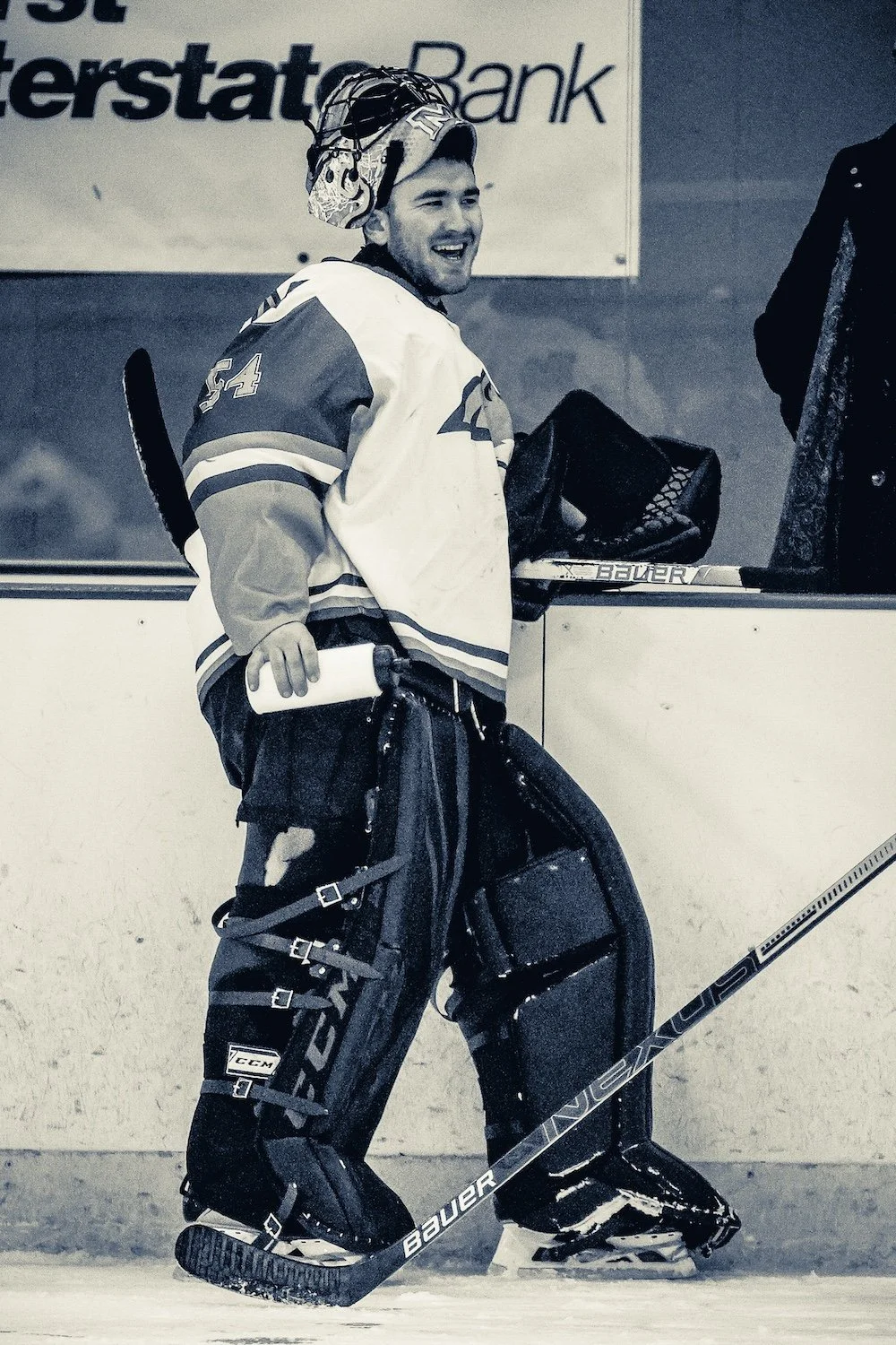 A hockey goalie standing on the ice in full gear, holding a hockey stick and a water bottle, with a smiling expression, near a rink barrier.
