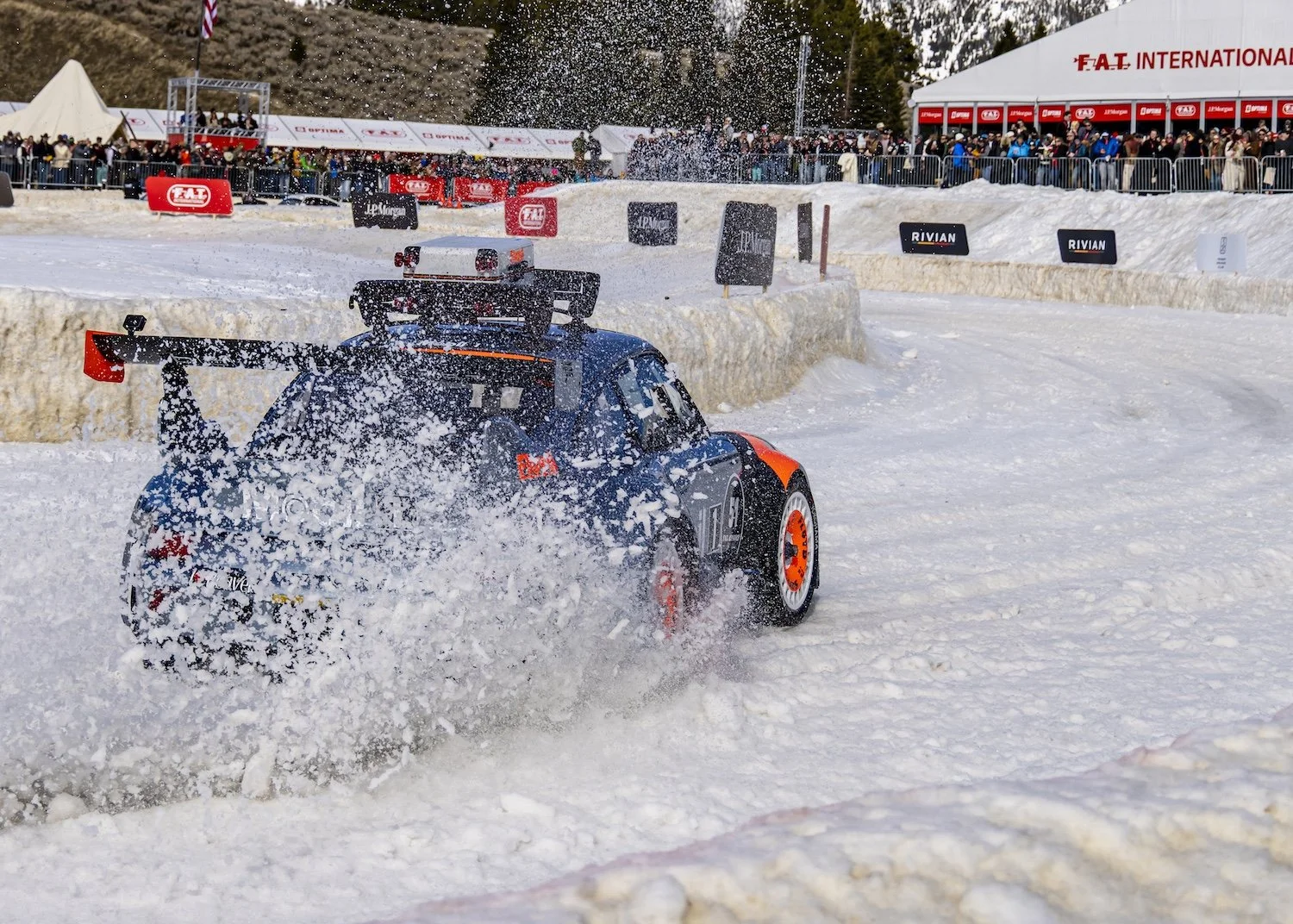 A rally car racing on snow, kicking up snow spray, with spectators and event tents in the background. F.A.T. ICE RACE 2026, Big Sky, Montana USA. Dan Lakatos Photo