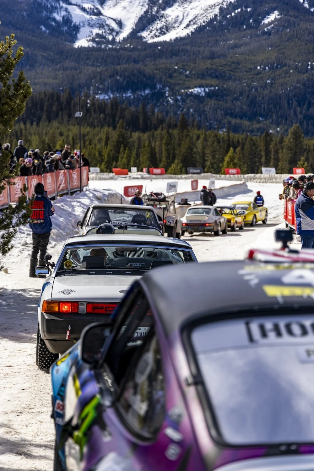 Vintage race cars parked on a snow-covered mountain road during a racing event, with spectators and marshals watching. F.A.T. ICE RACE 2026, Big Sky, Montana USA. Dan Lakatos Photo