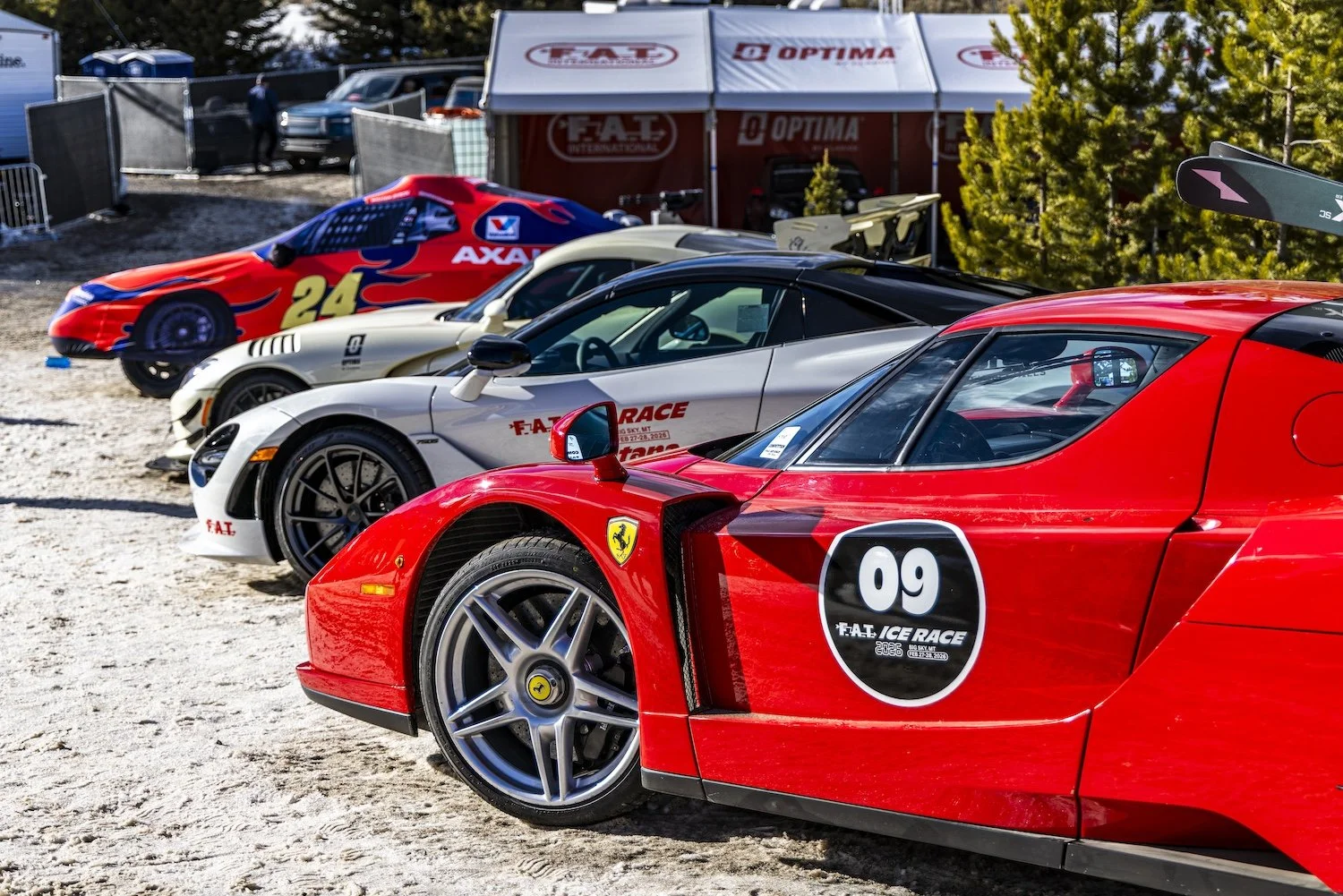Four race cars, including a red Ferrari with number 09, are parked on a dirt surface at a race event with tents and trees in the background. F.A.T. ICE RACE 2026, Big Sky, Montana USA. Dan Lakatos Photo