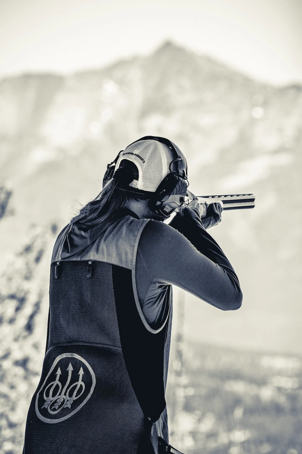 A person dressed in shooting gear aiming a shotgun outdoors with mountains in the background.