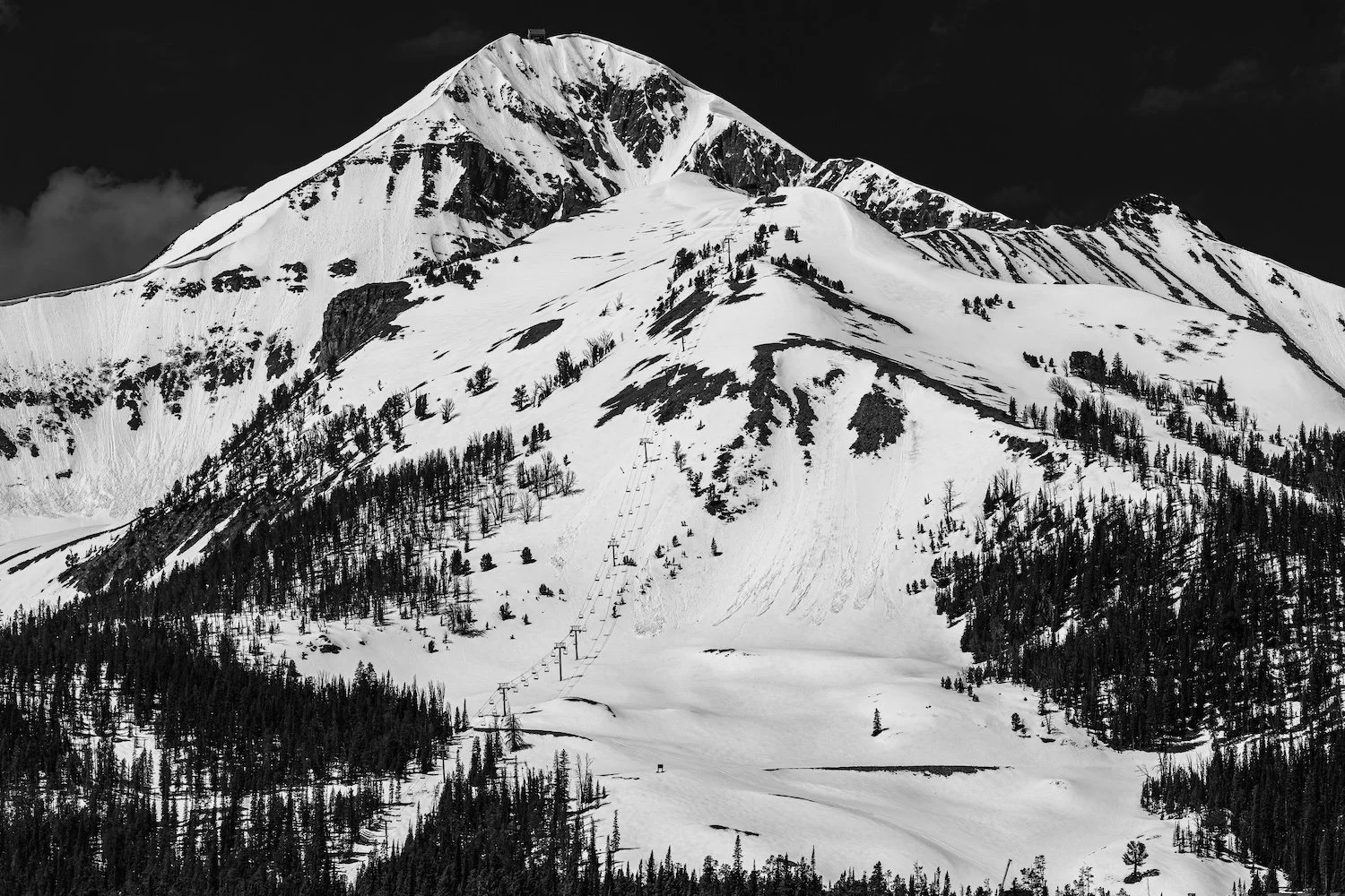 Snow-covered mountain with ski lift lines and trees at the base, under a dark sky.