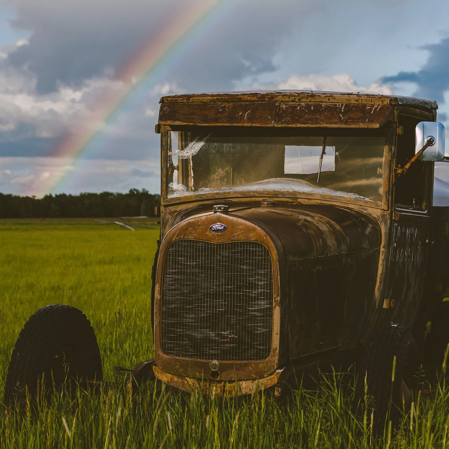 An old rusty Ford truck in a green field with a rainbow and dark clouds in the sky. Bozeman, Montana.