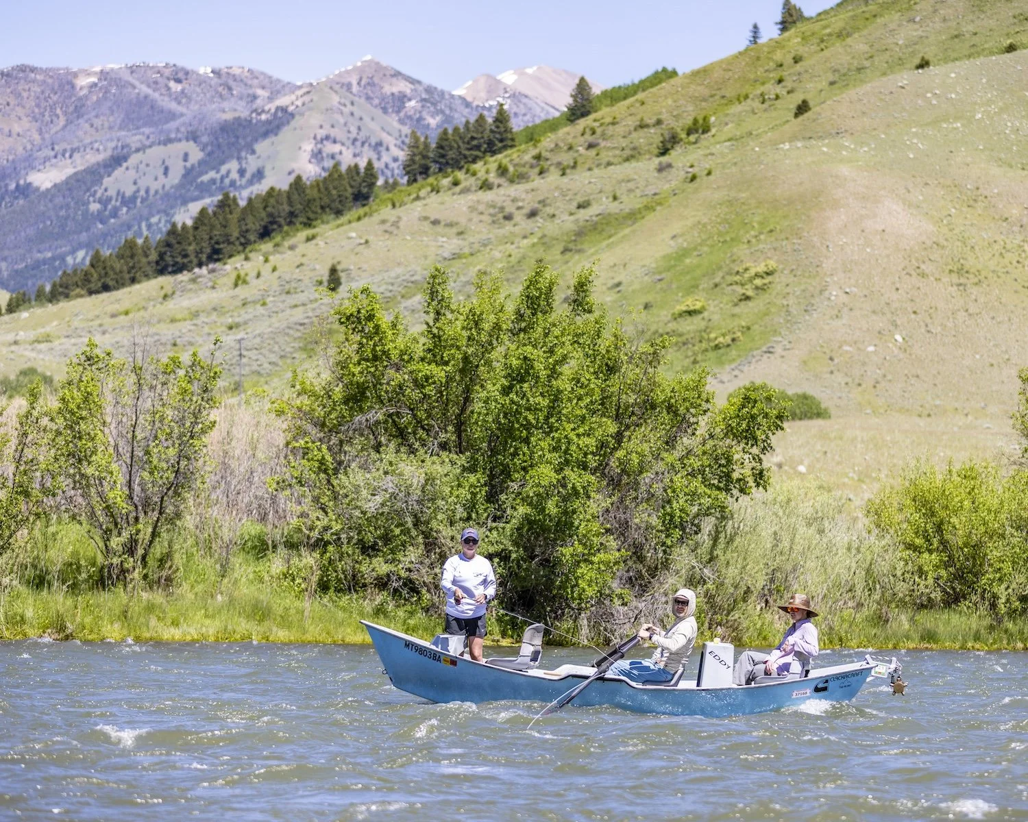 Three people fishing on a boat in a river surrounded by green trees, with mountains in the background on a sunny day. Madison River, Montana, USA