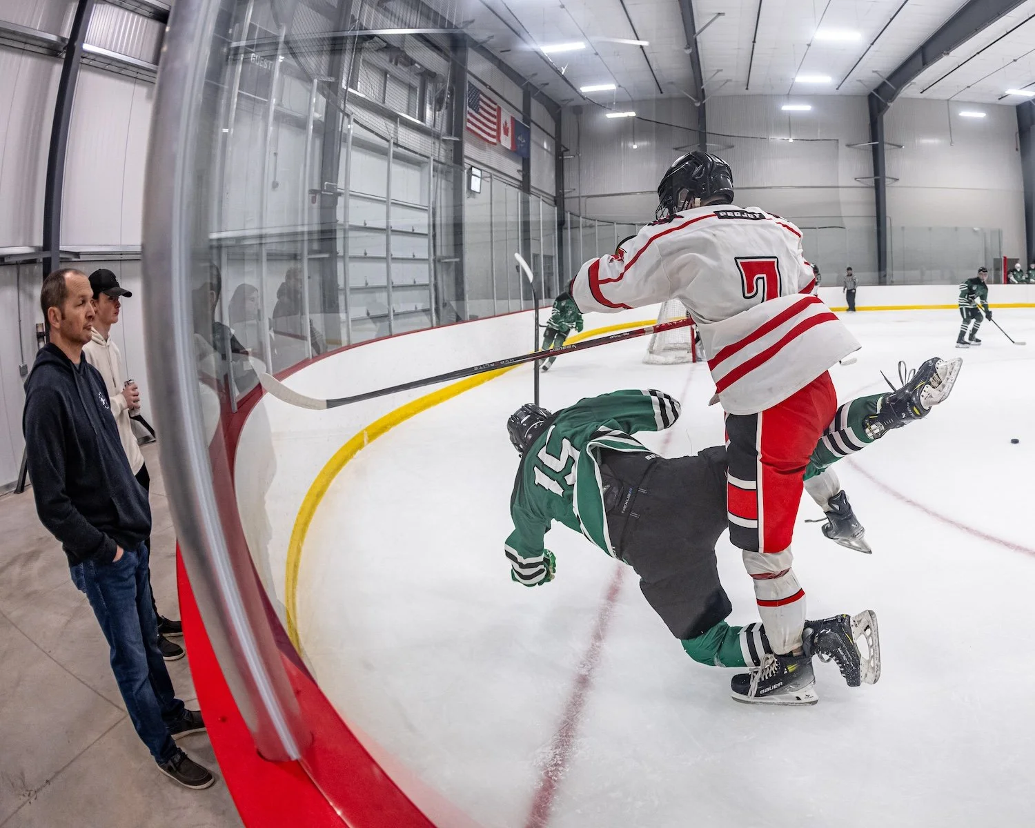 An ice hockey game showing a player in white and red jersey fighting with a player in green and black jersey near the ice rink boards. Three spectators are watching from outside the rink, and other players are visible in the background.