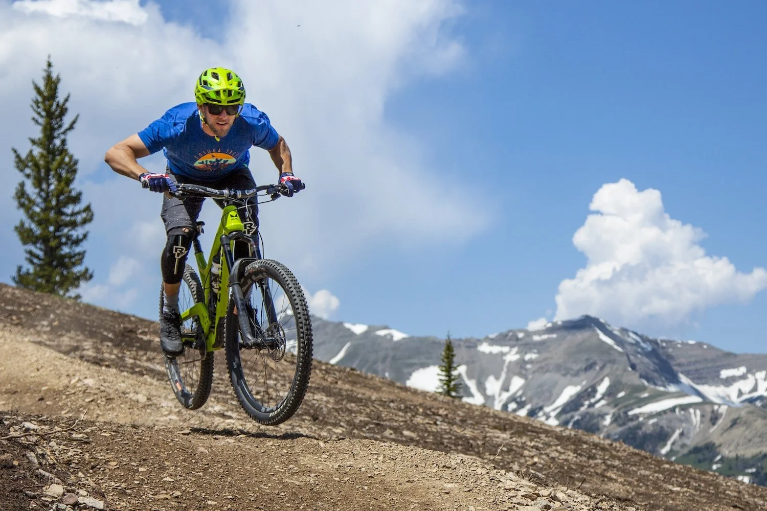 A mountain biker wearing a blue shirt, green helmet, and protective gear jumps off a dirt trail with snow-capped mountains and a partly cloudy sky in the background. Big Sky, MT, USA