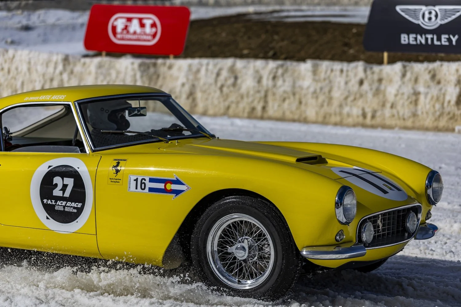 Yellow vintage Ferrari race car on a snowy track with racing decals and banners in the background. F.A.T. ICE RACE 2026, Big Sky, Montana USA. Dan Lakatos Photo