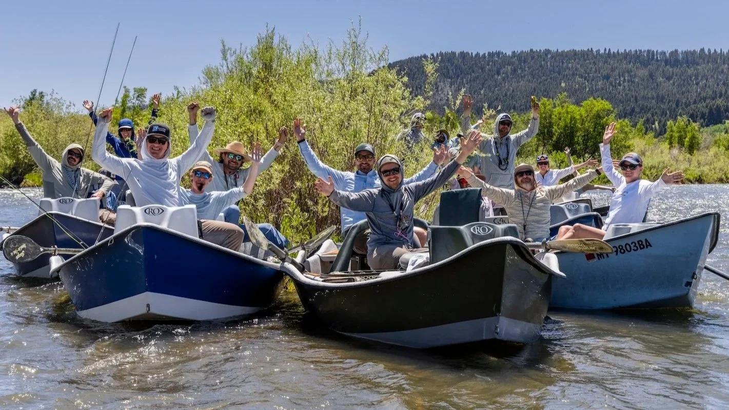 A group of people on two boats celebrating and smiling, with arms raised, on a river surrounded by green trees and mountains in the background.