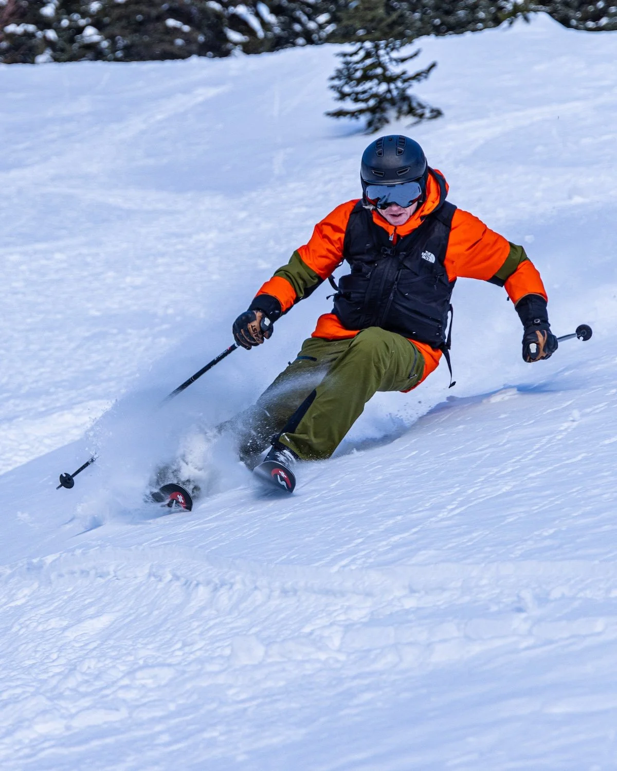 A skier in an orange jacket, green pants, and black helmet skiing down a snowy slope.