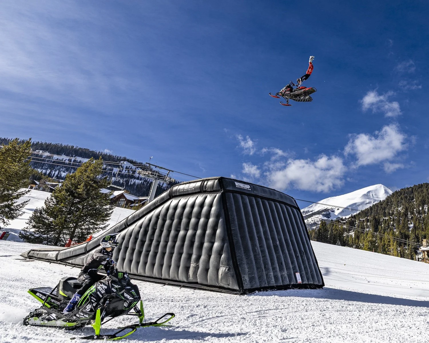 A person on a snowmobile at a ski resort, with a skier in mid-air performing a jump above a large inflatable ramp, snow-covered mountains, trees, and ski lift in the background under a blue sky with some clouds. Freestyle snowmobile show, Kieth Sayer