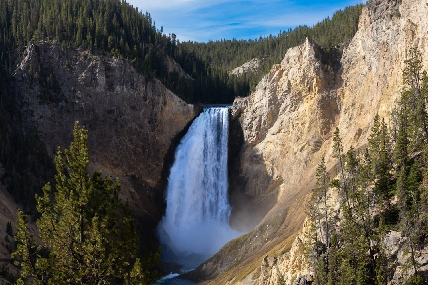 A tall waterfall cascading down a rocky cliff surrounded by green pine trees in a canyon with a blue sky overhead.