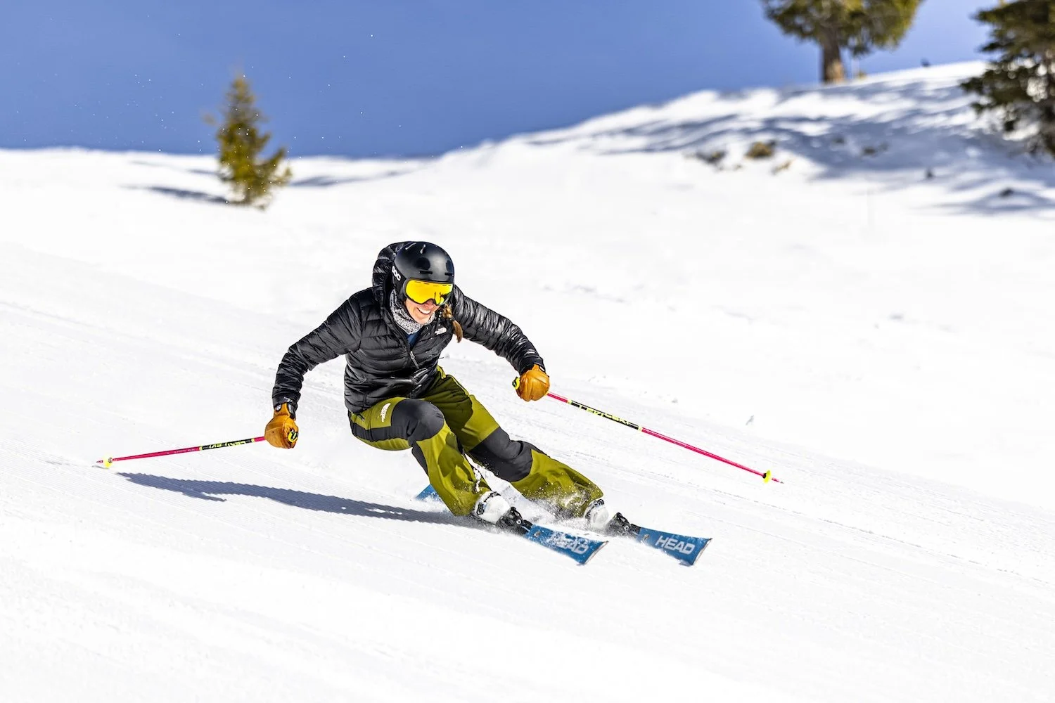 Person wearing black helmet and goggles skiing downhill on snow, dressed in black jacket and yellow-green pants, with poles, in a snowy landscape with trees