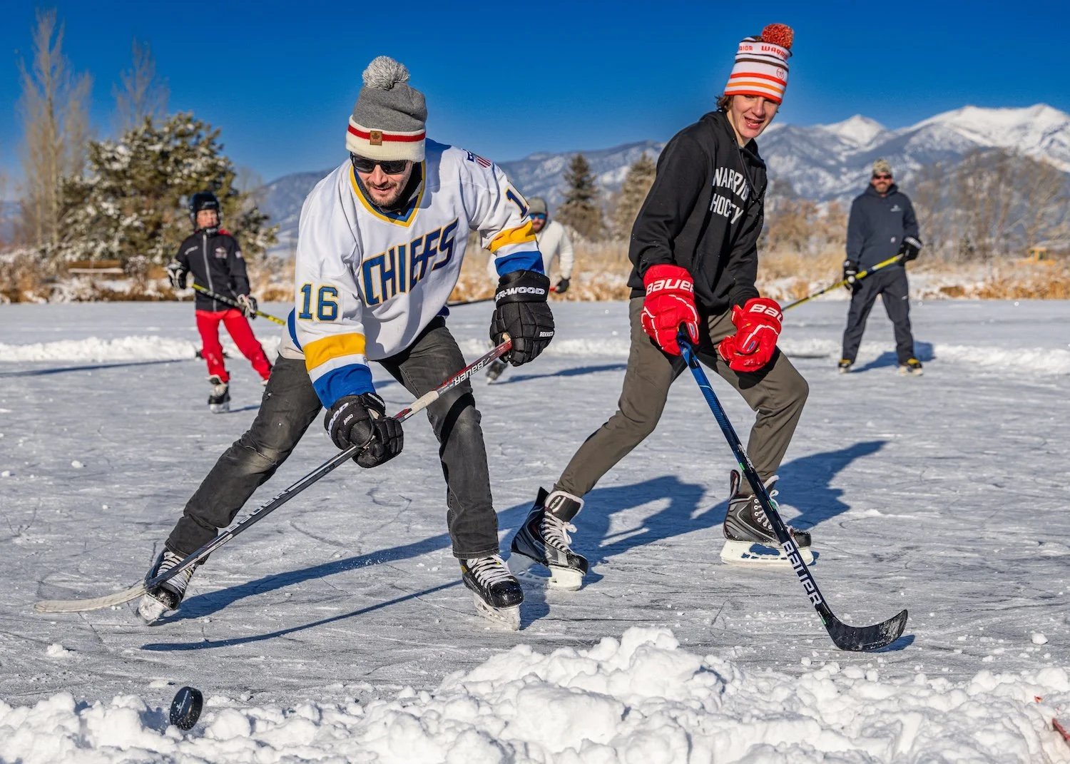 People playing hockey on an outdoor snowy rink with mountains in the background.
