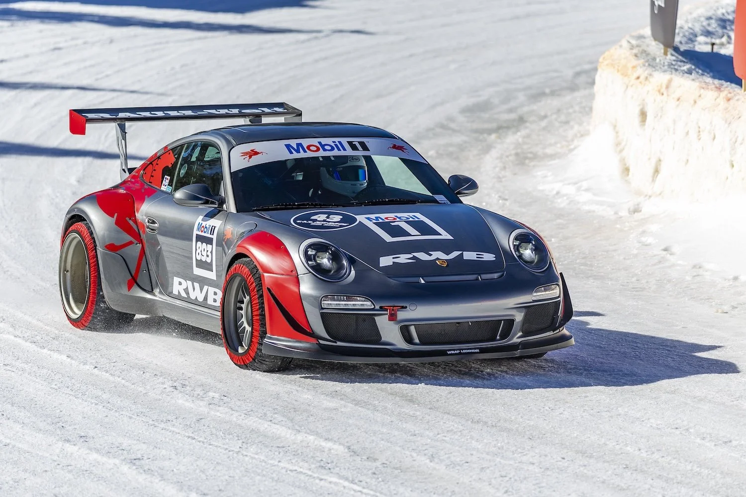 A race car driving on snow-covered track with snow banks on the side. The car is gray with red accents and various sponsor logos, including Mobil 1 and RWB. It has a large rear wing and winter tires with red sidewalls. F.A.T. ICE RACE 2026, Big Sky, 