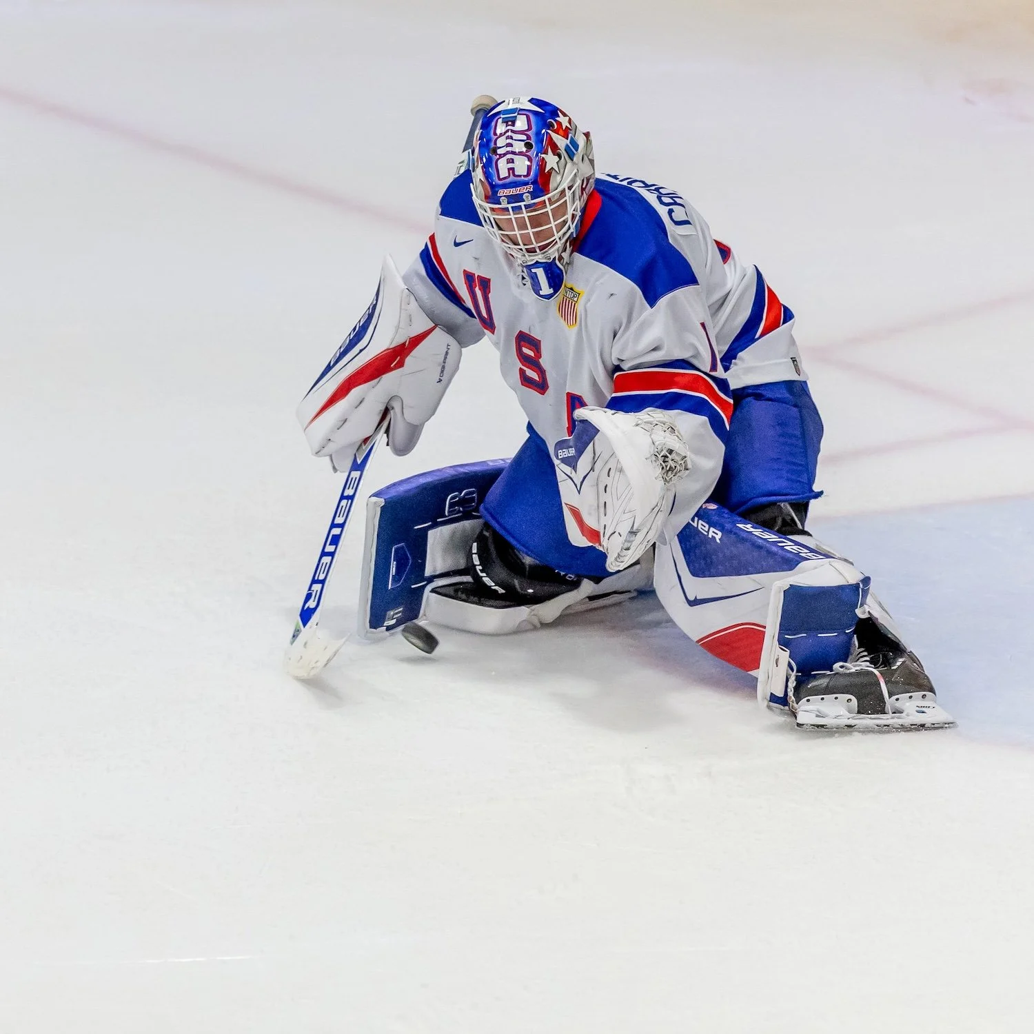 An ice hockey goaltender in white, red, and blue uniform crouching on the ice with a hockey stick, wearing a helmet and protective gear.