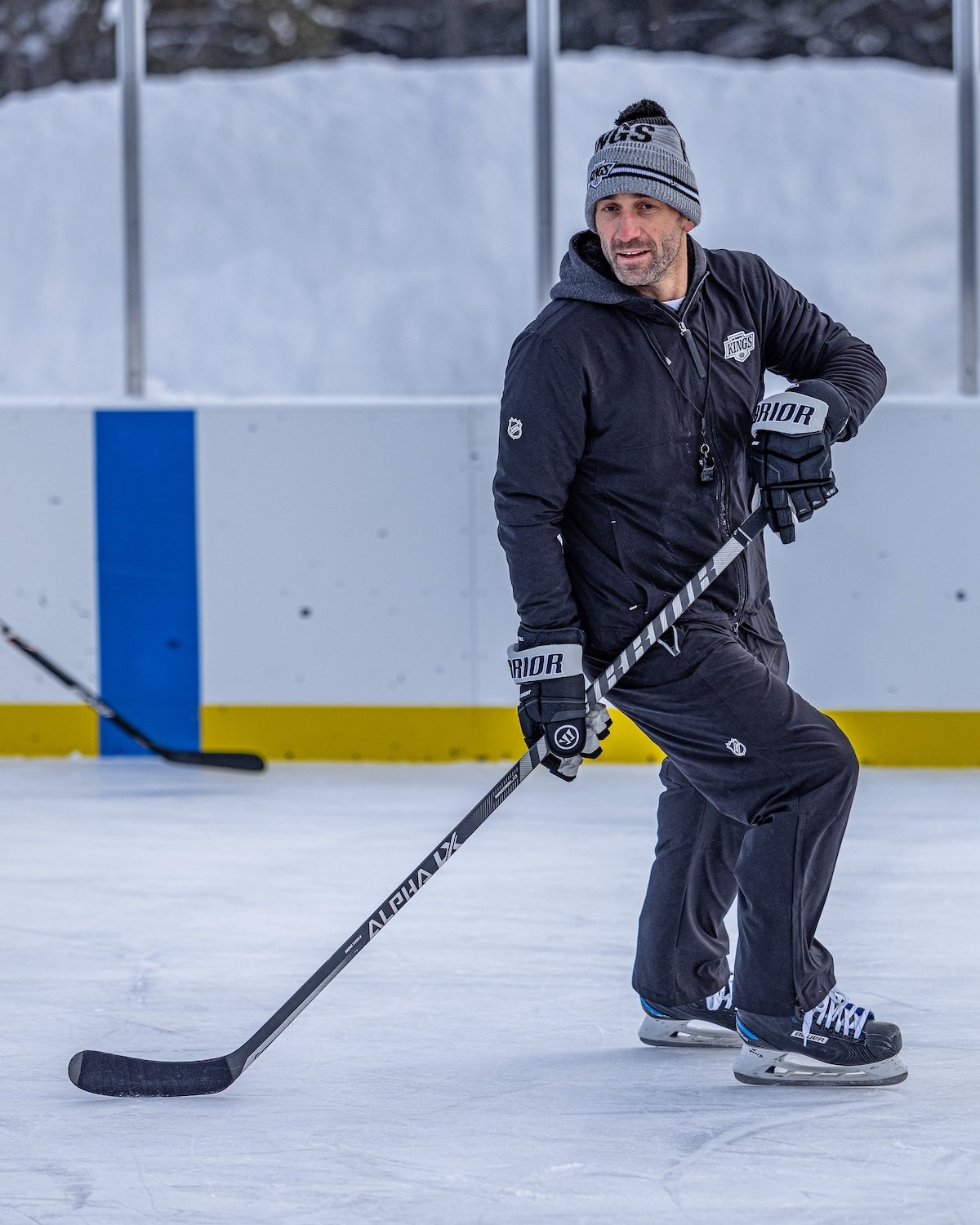 Man playing hockey on ice rink, wearing gray beanie, black jacket, black pants, and skates, holding a hockey stick.