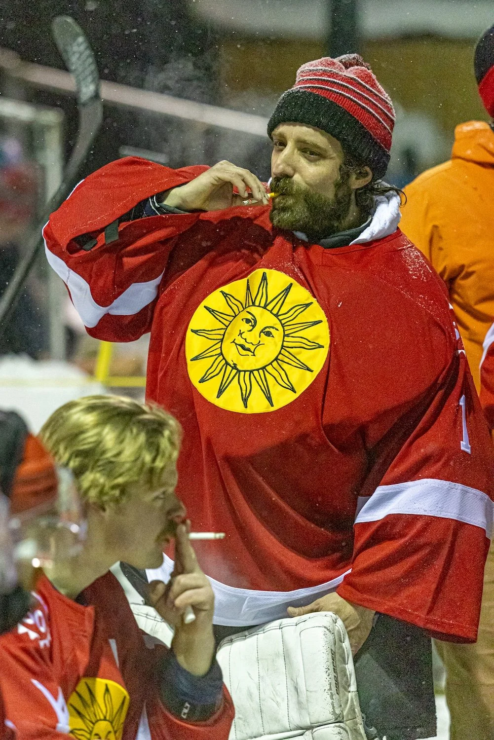 A man in a red hockey jersey and winter hat drinking from a cigarette holder, with a woman in a similar jersey and a hockey mask standing nearby.