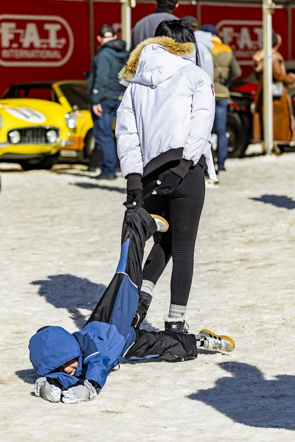 Child lying on snow with woman holding their ankle during a winter outdoor event. People and vintage cars are in the background. F.A.T. ICE RACE 2026, Big Sky, Montana USA. Dan Lakatos Photo