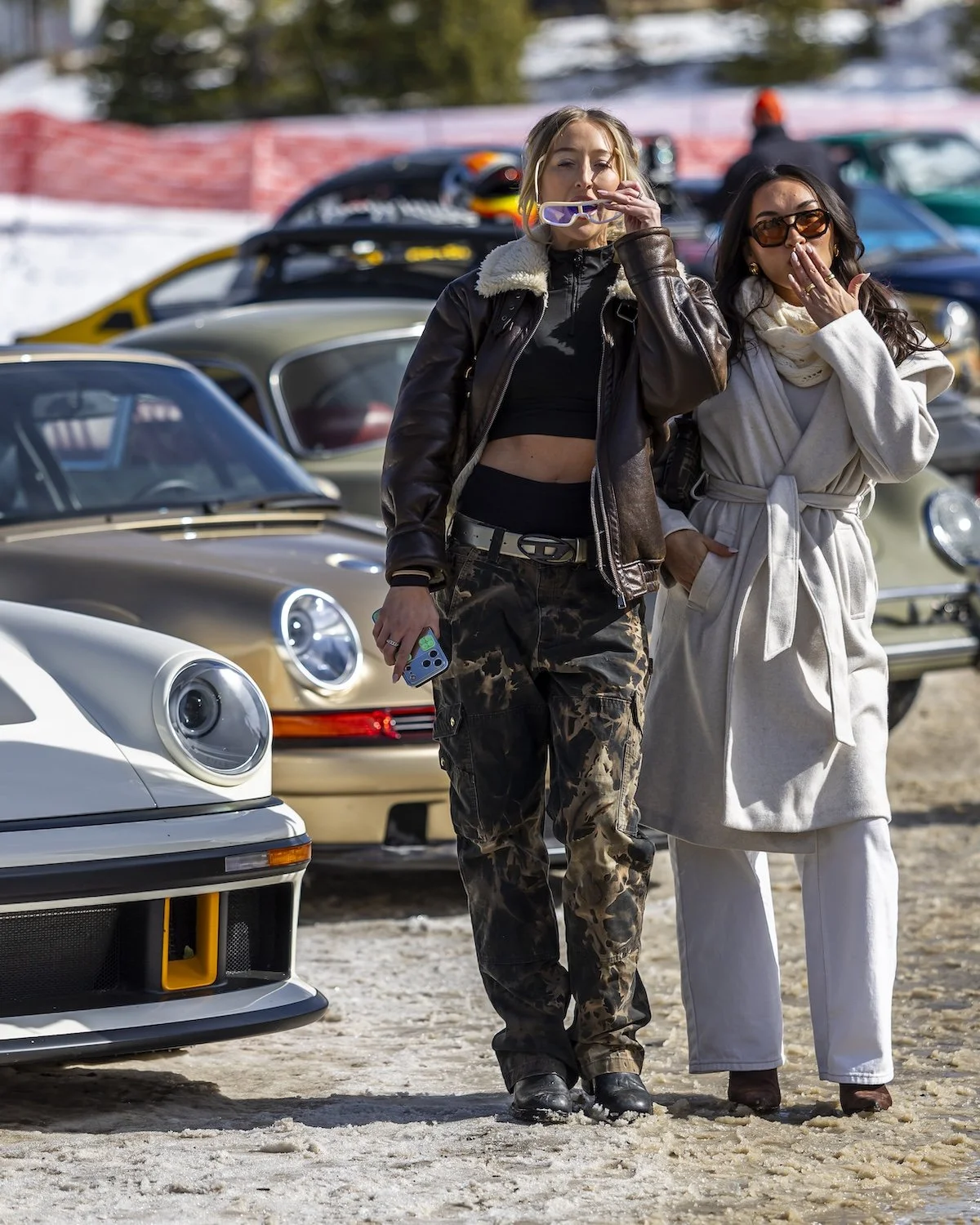 Two women standing in front of a collection of vintage cars, with one holding a smartphone and the other covering her mouth as they look at the camera F.A.T. ICE RACE 2026, Big Sky, Montana USA. Dan Lakatos Photo