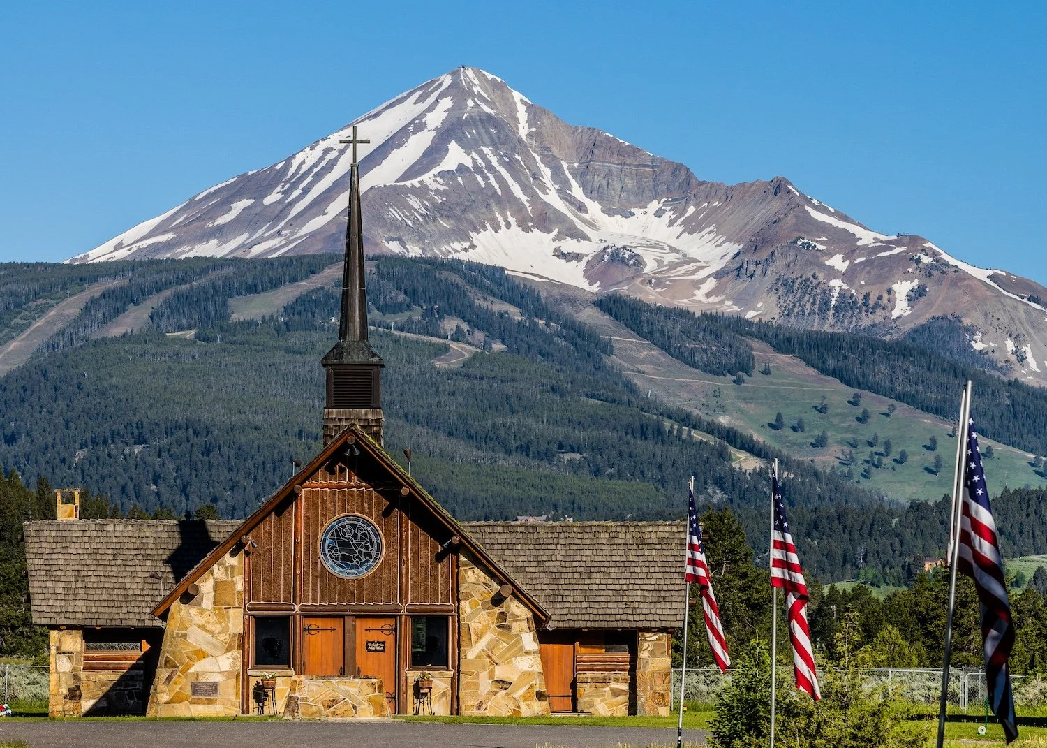 Soldier's Chapel with a stone facade and wooden doors, surrounded by American flags, with a snow-capped mountain in the background under a clear blue sky. Big Sky, Montana