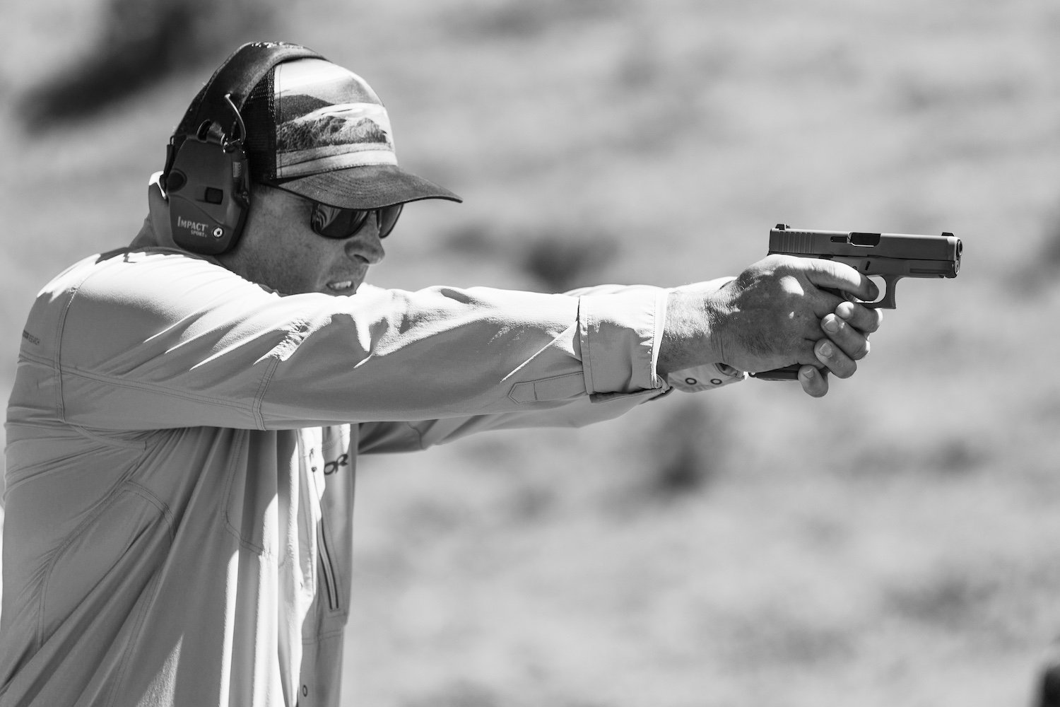 A man wearing sunglasses, a cap, and hearing protection, aiming a handgun during a shooting session outdoors. Montana, USA
