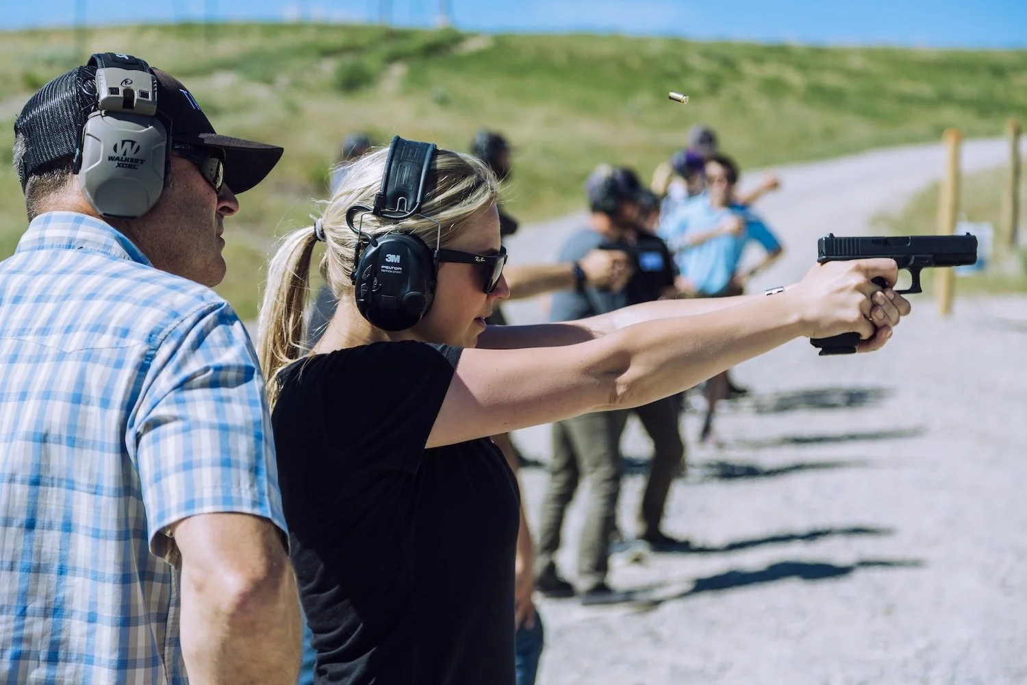 A group of people at a shooting range outdoors, with a woman aiming a handgun, wearing protective ear and eye gear, and others standing behind her in the background. Montana, USA