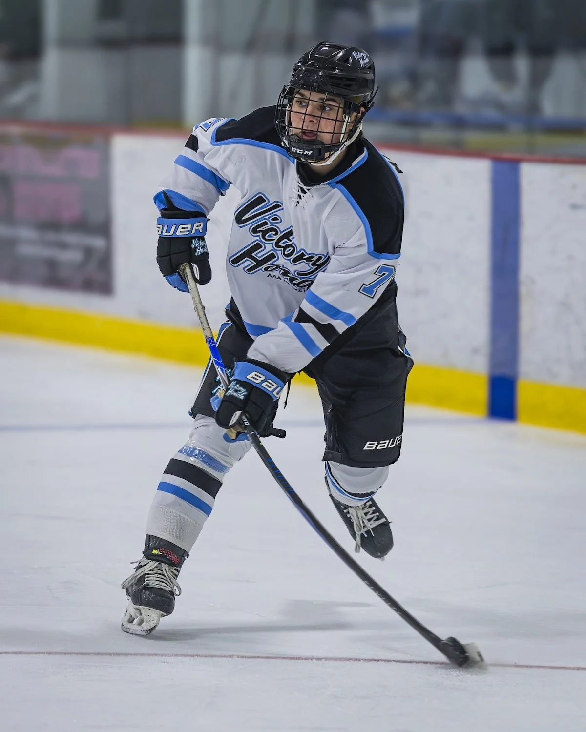 A male ice hockey player wearing a black helmet, white jersey with blue and black accents, and black shorts, skating on the ice with a hockey stick.