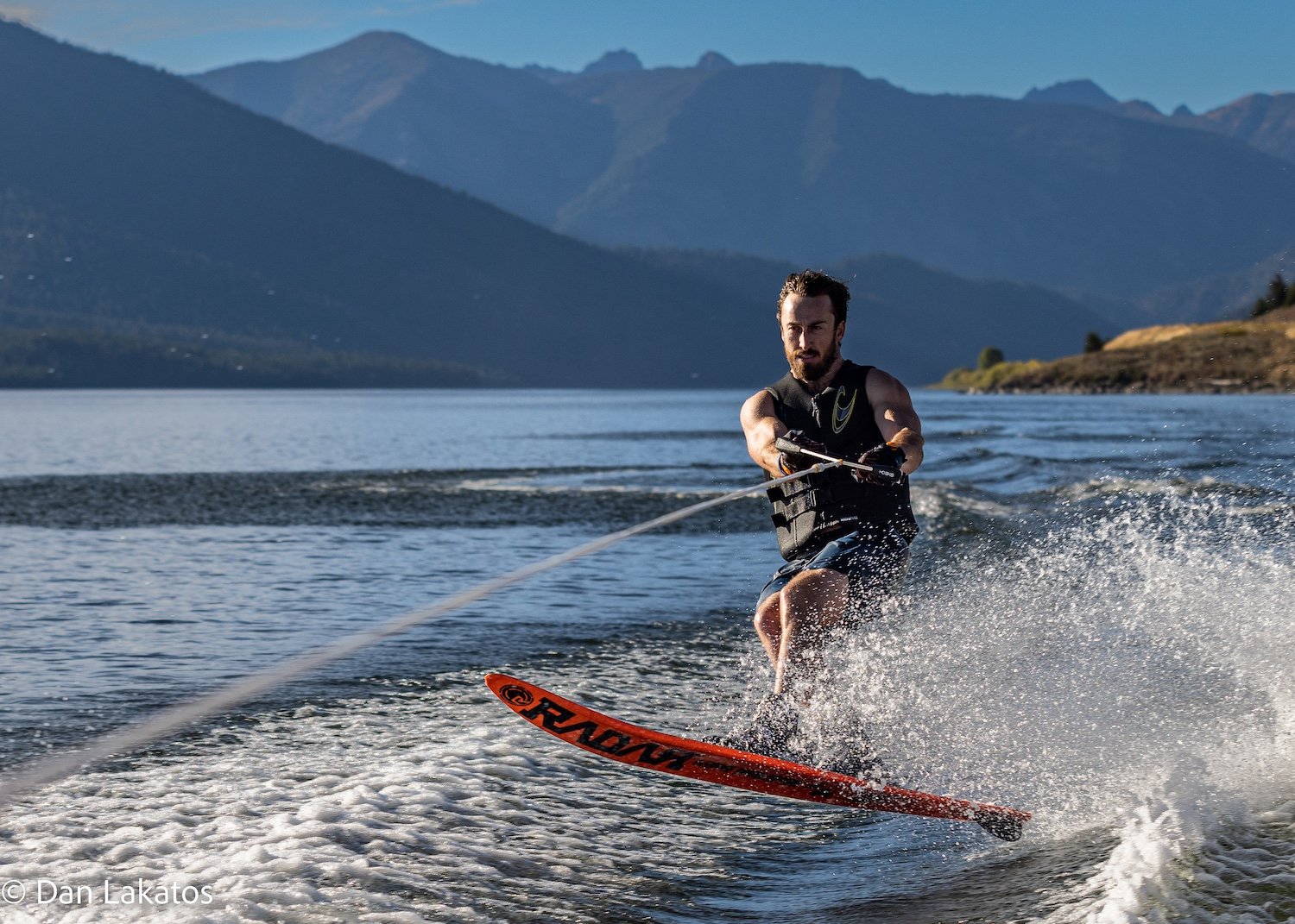A man waterskis on a lake with mountains in the background, wearing a life vest and holding a tow rope. Hebgen Lake, Montana, USA