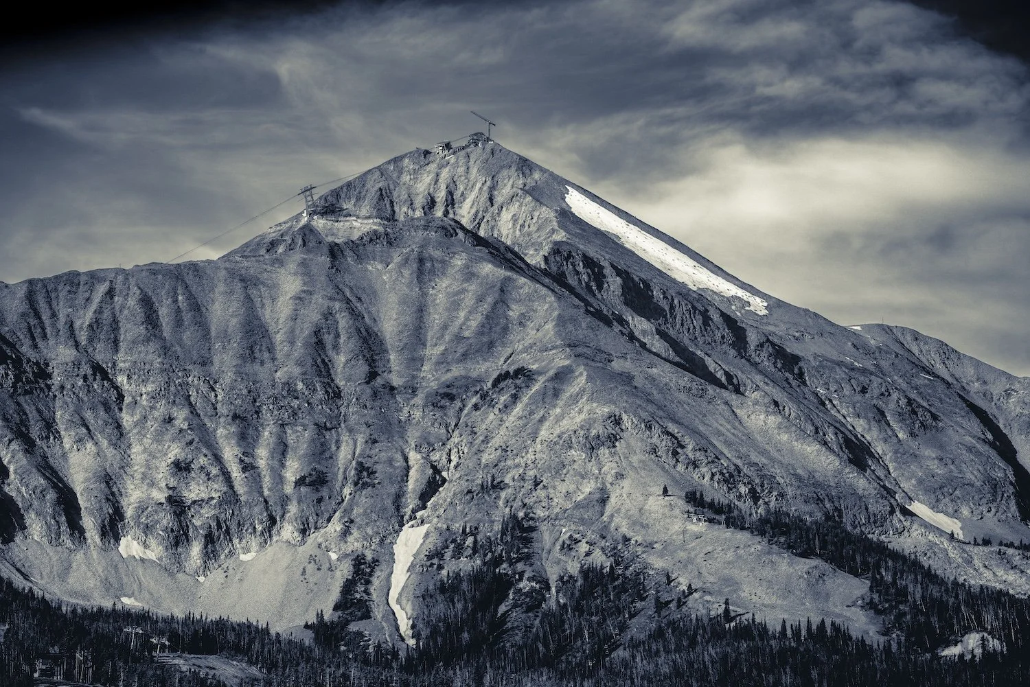A mountain with a snowy peak and rugged, rocky slopes under a cloudy sky, with ski lift cables and a ski area at the base. Lone Peak, Moonlight Basin, Big Sky, Montana, USA