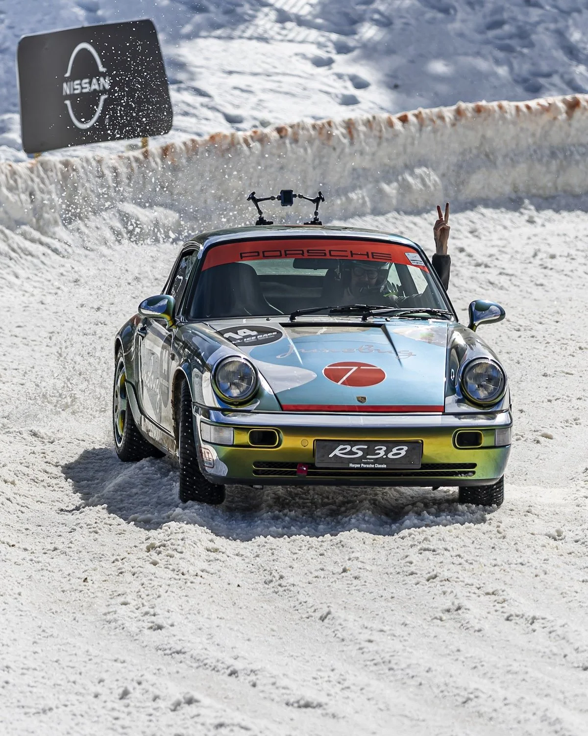 A vintage Porsche RS 3.8 race car on snow, with a person inside making a peace sign and a hand waving. There is a Nissan sign in the background. F.A.T. ICE RACE 2026, Big Sky, Montana USA. Dan Lakatos Photo