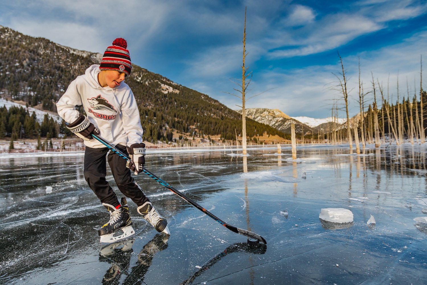 A young person ice skating on a frozen lake with mountains and trees in the background.