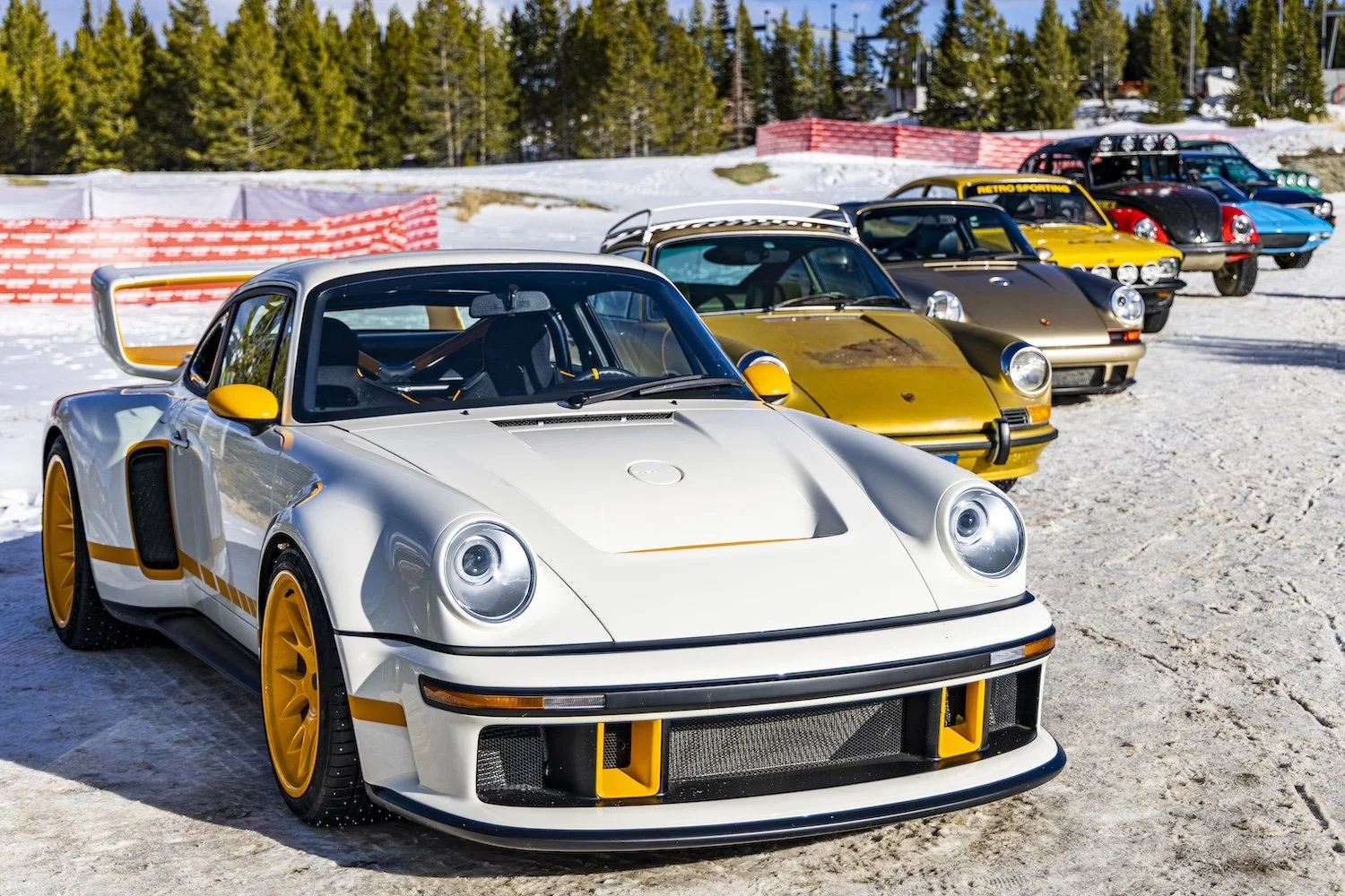 A lineup of colorful Porsche sports cars on a snowy outdoor track, featuring a white Porsche 911 with yellow accents in the foreground and other vintage models in the background. F.A.T. ICE RACE 2026, Big Sky, Montana USA. Dan Lakatos Photo