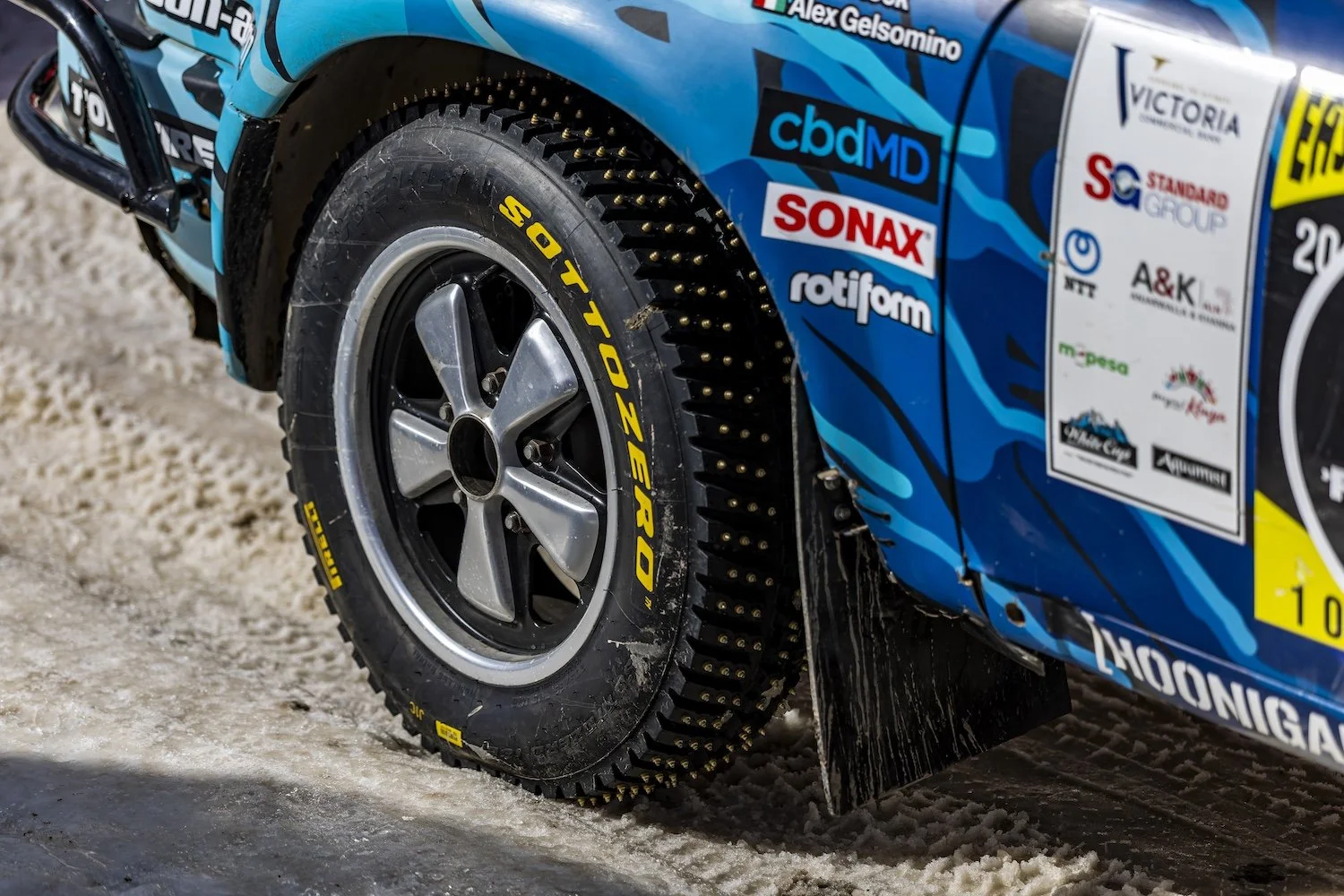 Close-up of a race car's rear wheel with a Pirelli Sottozero tire, large studded tire spikes, and blue racing livery with sponsor decals on a snowy surface. F.A.T. ICE RACE 2026, Big Sky, Montana USA. Dan Lakatos Photo