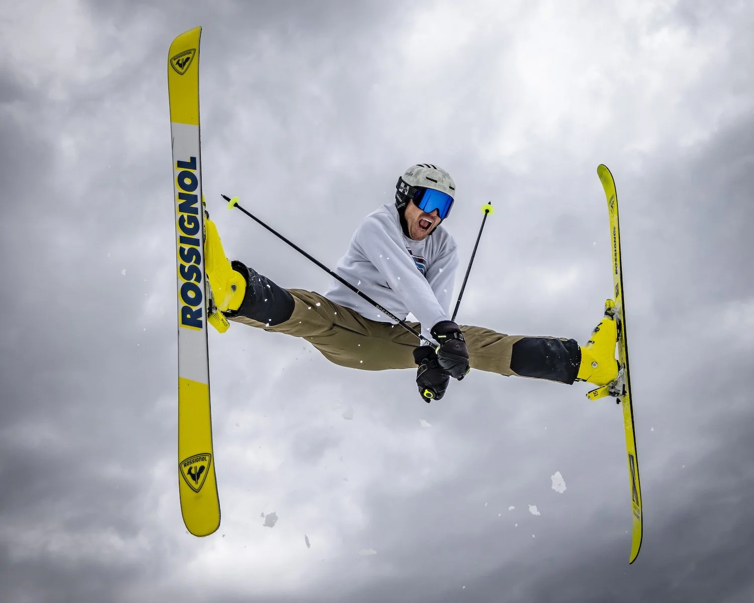 Skier performing a stunt in mid-air with legs spread wide, wearing a white jacket, tan pants, yellow skis, and a helmet with goggles against a cloudy sky.