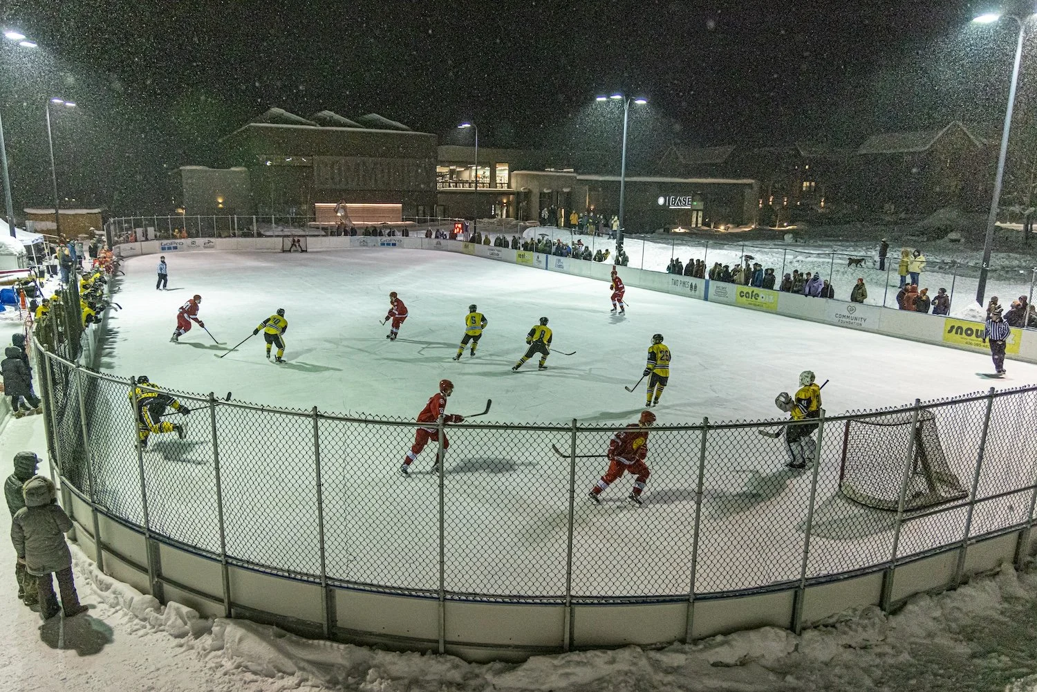 An ice hockey game taking place at an outdoor rink at night, with players in red and yellow jerseys, surrounded by spectators and snow.
