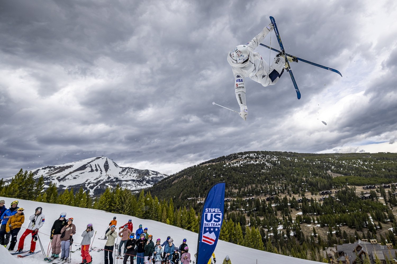 A skier in mid-air executing a stunt during a skiing event with spectators watching below, set in a mountainous area with snow, trees, and cloudy sky.