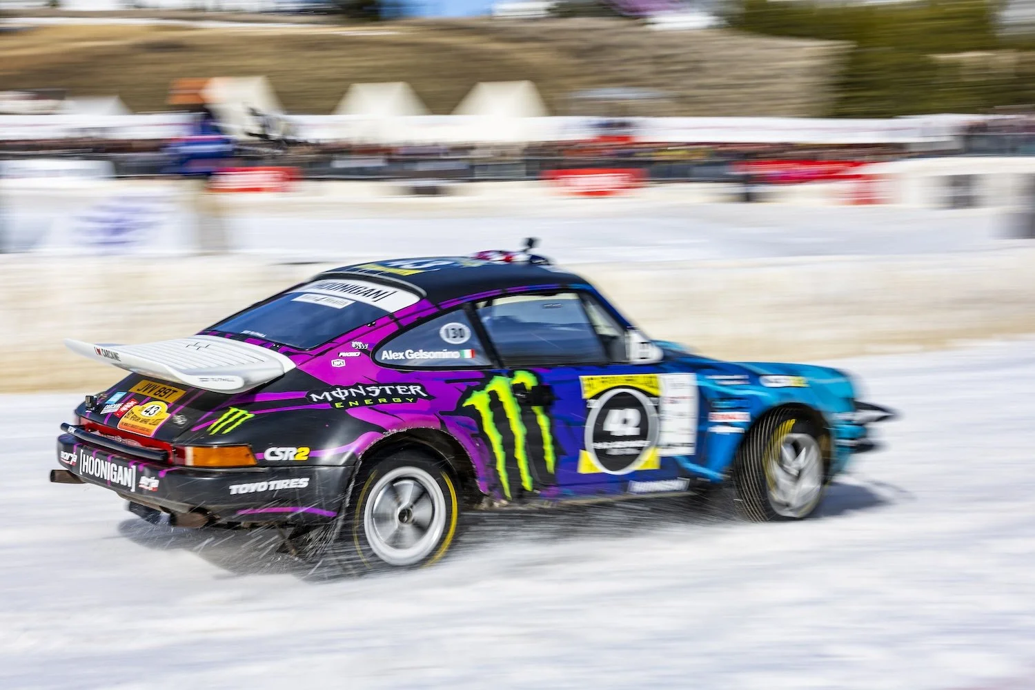 A Porsche race car speeding on a snow-covered track with colorful sponsor logos and a blurred background indicating motion. F.A.T. ICE RACE 2026, Big Sky, Montana USA. Dan Lakatos Photo