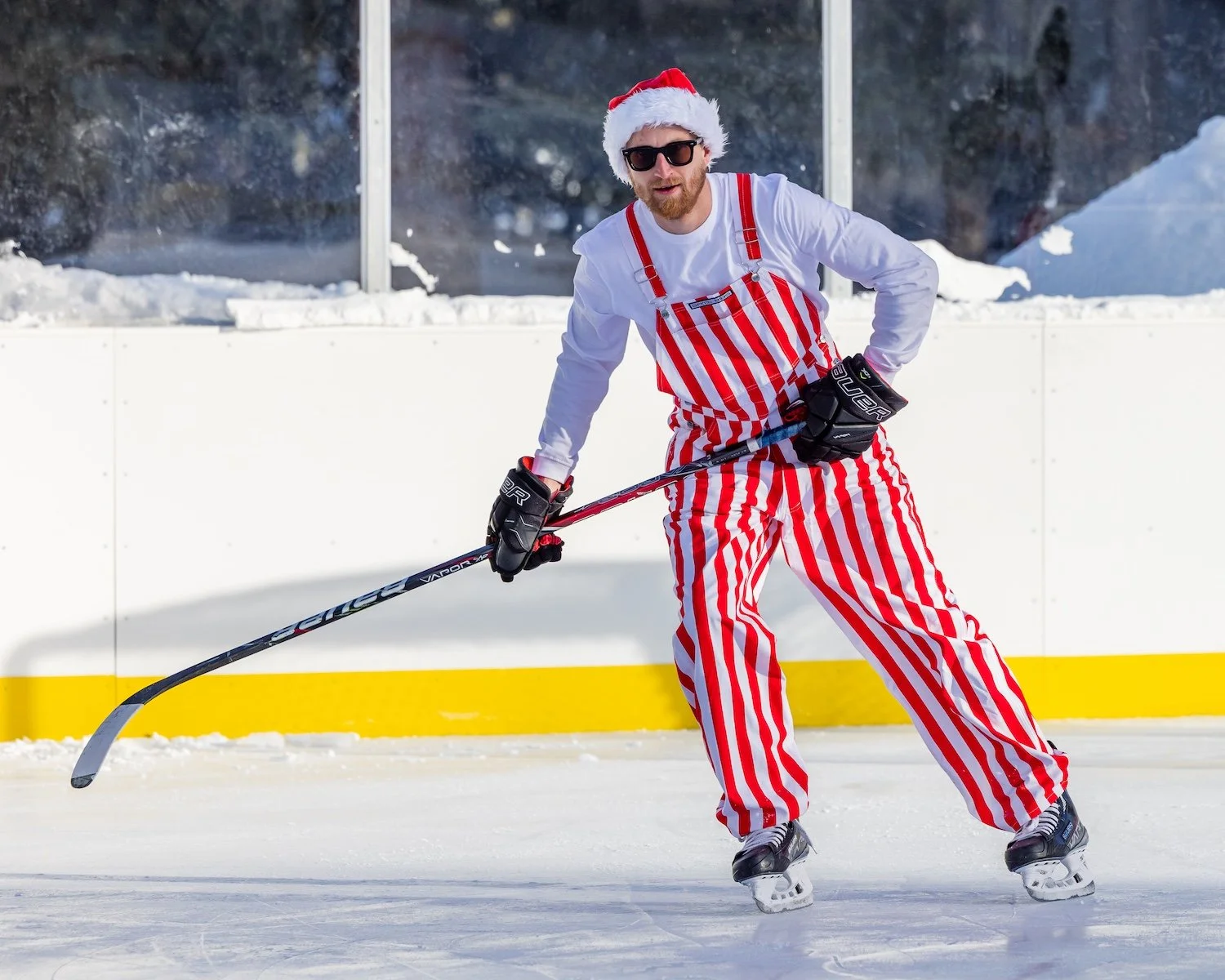 A man dressed in a Santa hat, sunglasses, and red-and-white striped pants and apron, playing ice hockey on an outdoor rink.