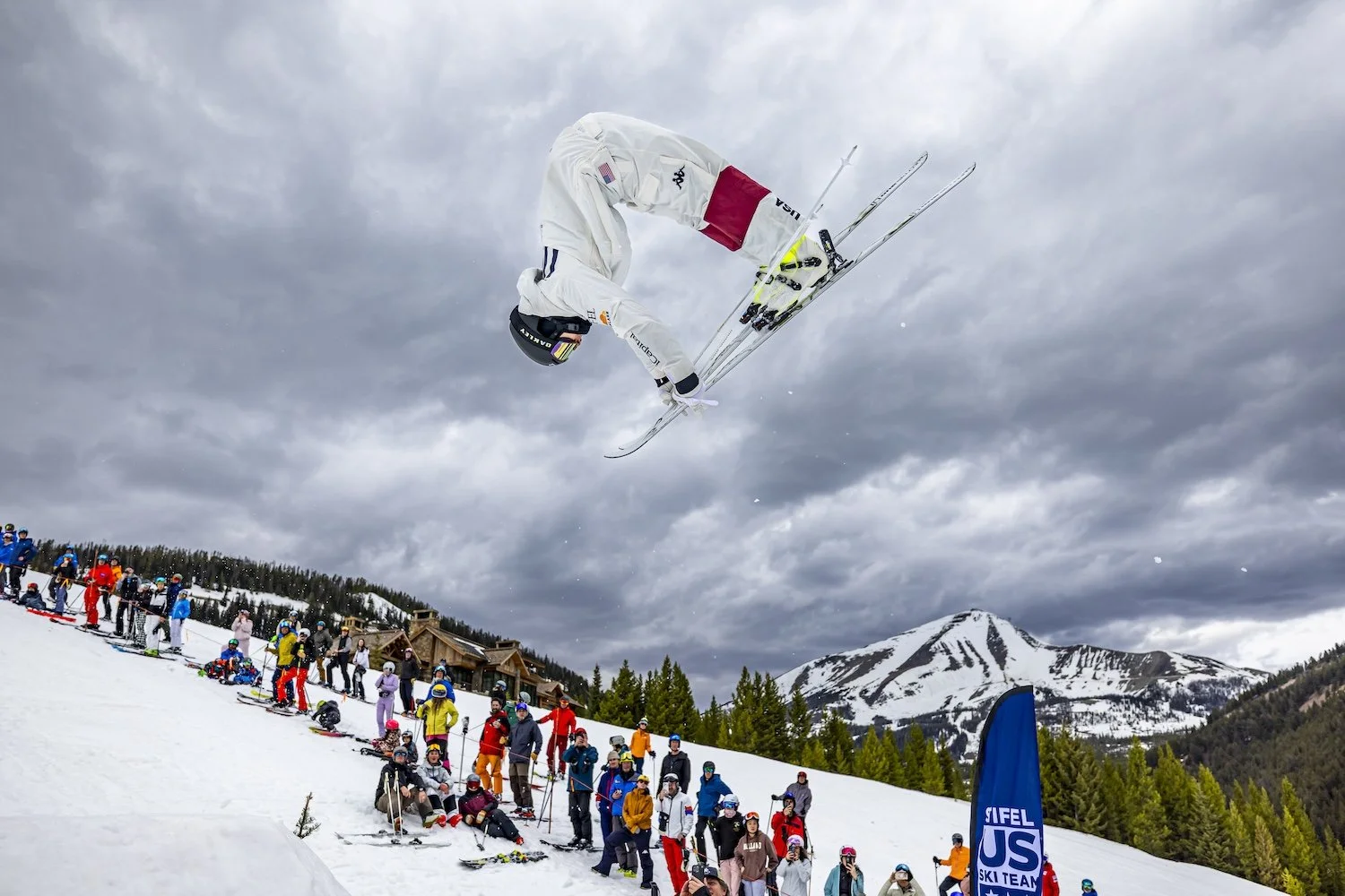 A skier in a white outfit and helmet performs an aerial trick in mid-air during a ski event on a snowy mountain slope, with spectators watching below under a cloudy sky.