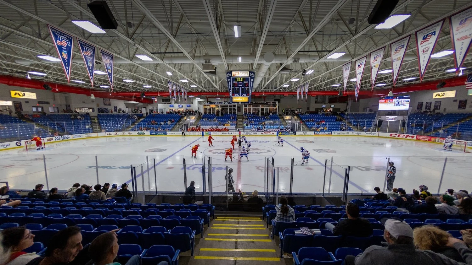 Indoor ice hockey rink with players in red and white jerseys, spectators in blue seats, and banners hanging from ceiling, with a scoreboard showing 00:00.