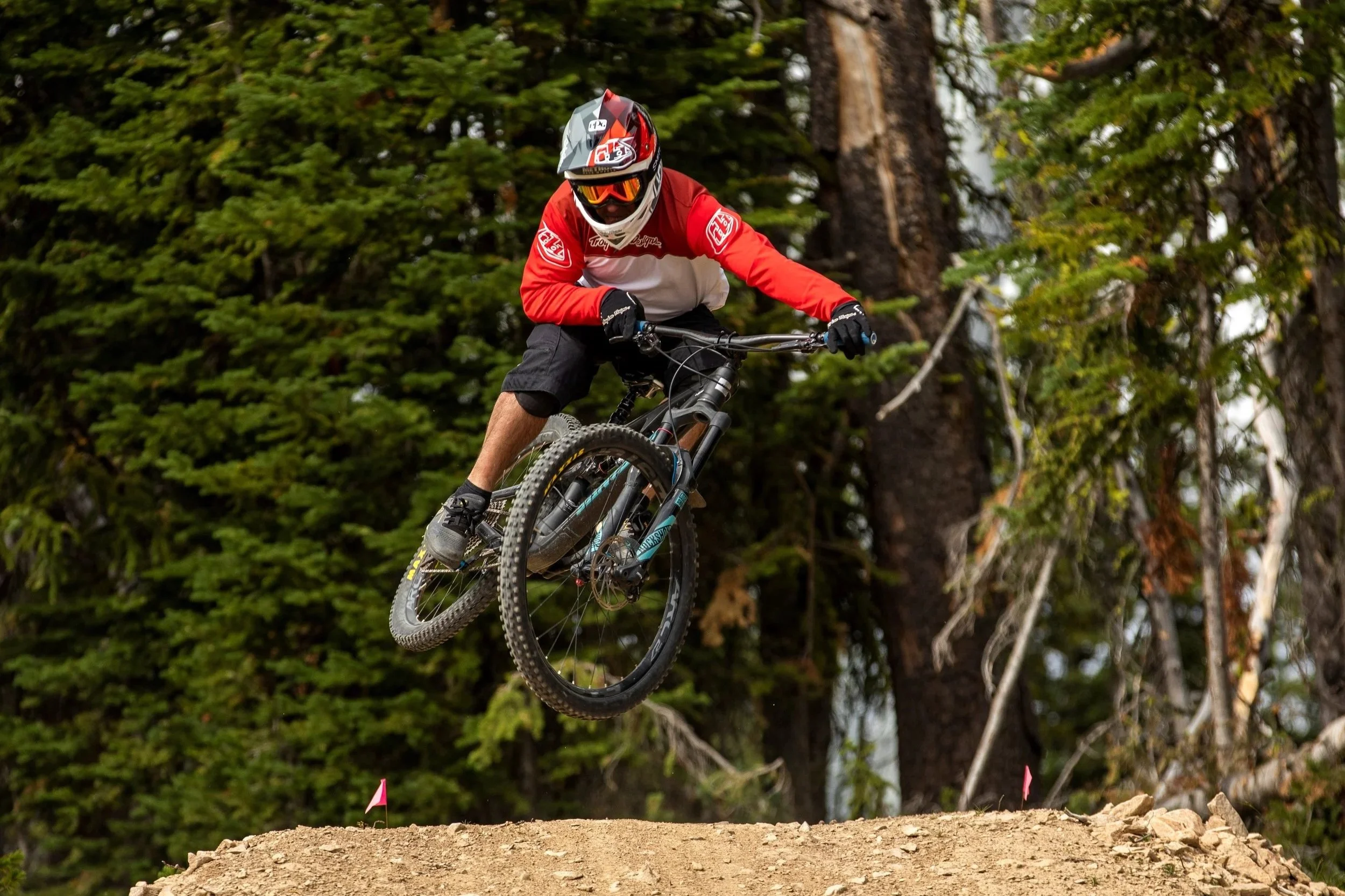 Mountain biker wearing a red and white jersey, black shorts, and full helmet riding a black mountain bike on a dirt trail in a forested area.