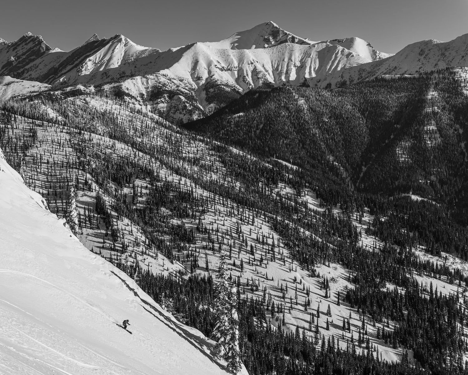 A skier skiing downhill on a snow-covered mountain slope with a backdrop of snow-capped mountain peaks and dense forests.