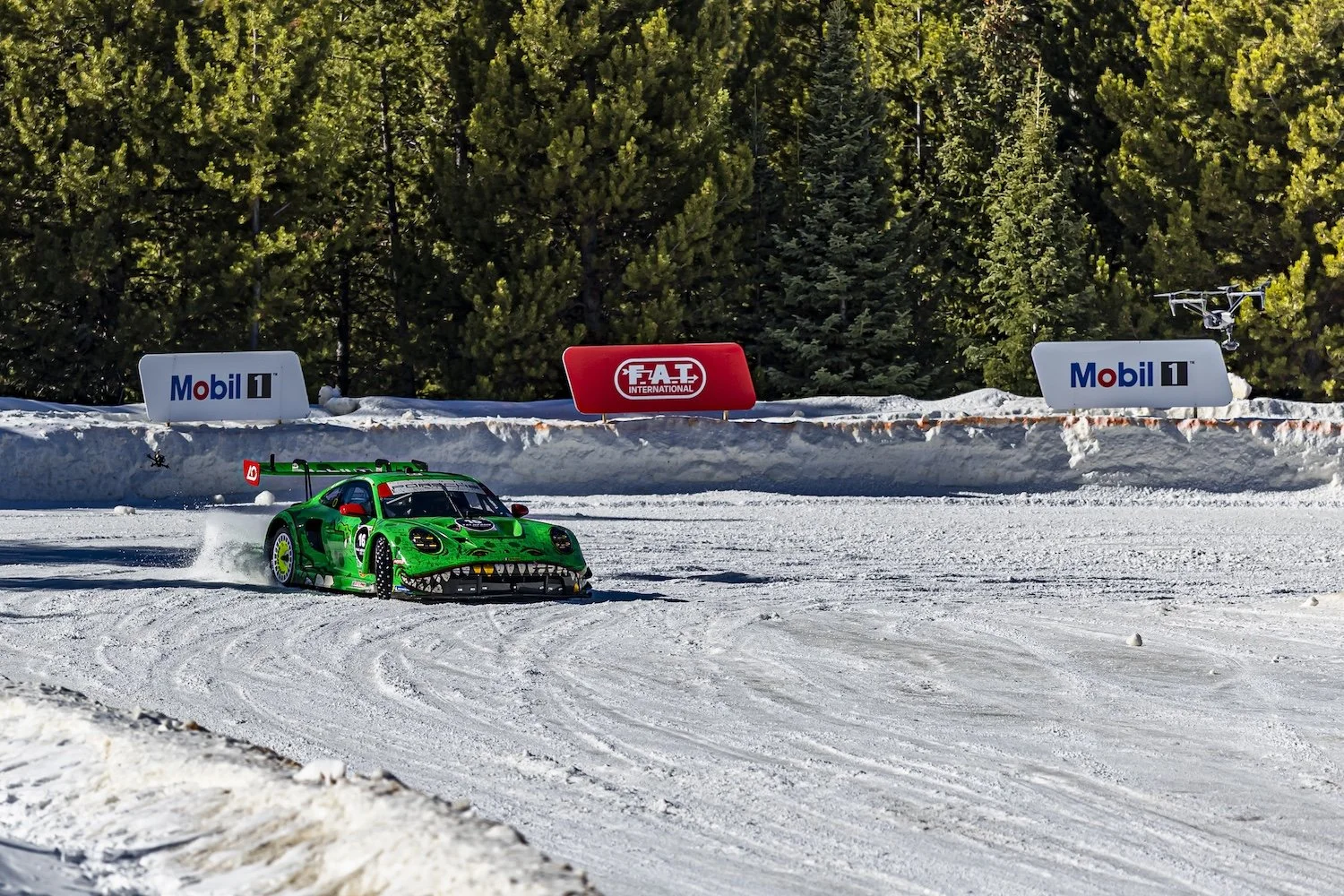 Green race car with shark teeth design driving on snow-covered race track with snow spray behind, in front of evergreen trees, advertising banners for Mobil 1 and F.A.T. International in the background. F.A.T. ICE RACE 2026, Big Sky, Montana USA. Dan