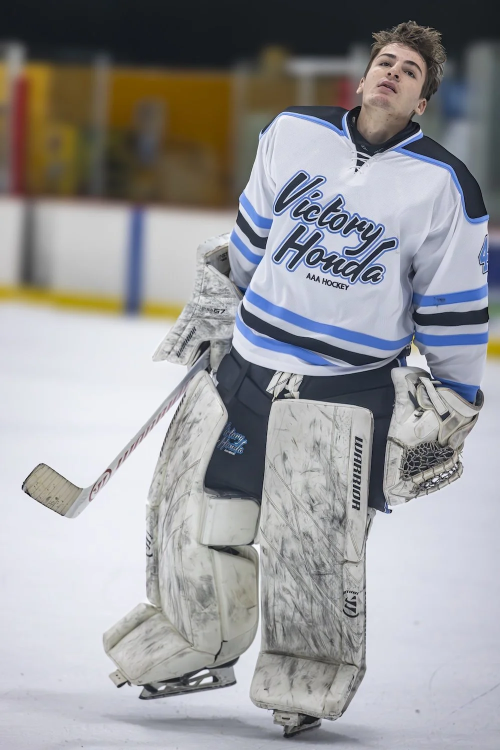 Ice hockey goalie in a white and blue jersey with 'Victory Honda AAA Hockey' logo, standing on ice rink with gloves and pads, holding a hockey stick, looking up with a slightly distressed expression.