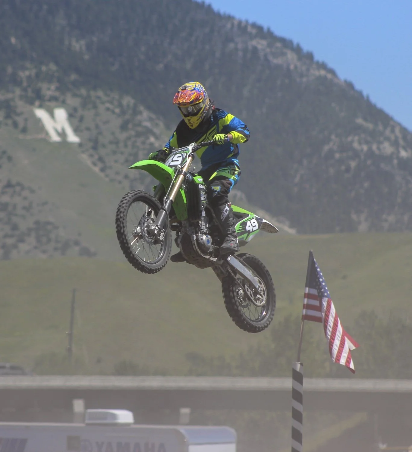 A motocross rider wearing a blue and yellow outfit and a colorful helmet is airborne on a green dirt bike over a dirt track, with American flags in the foreground and mountains in the background. Bozeman, Montana, USA