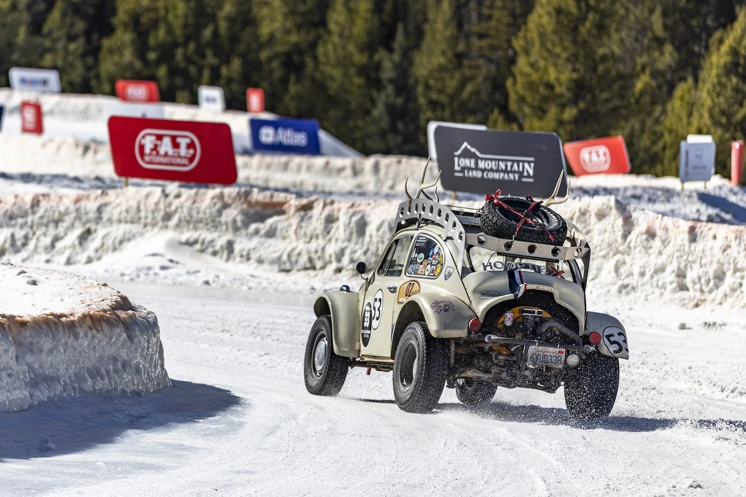 A vintage off-road vehicle driving on a snow-covered track during a race, with snow banks on the side and banners for various sponsors and brands in the background. F.A.T. ICE RACE 2026, Big Sky, Montana USA. Dan Lakatos Photo