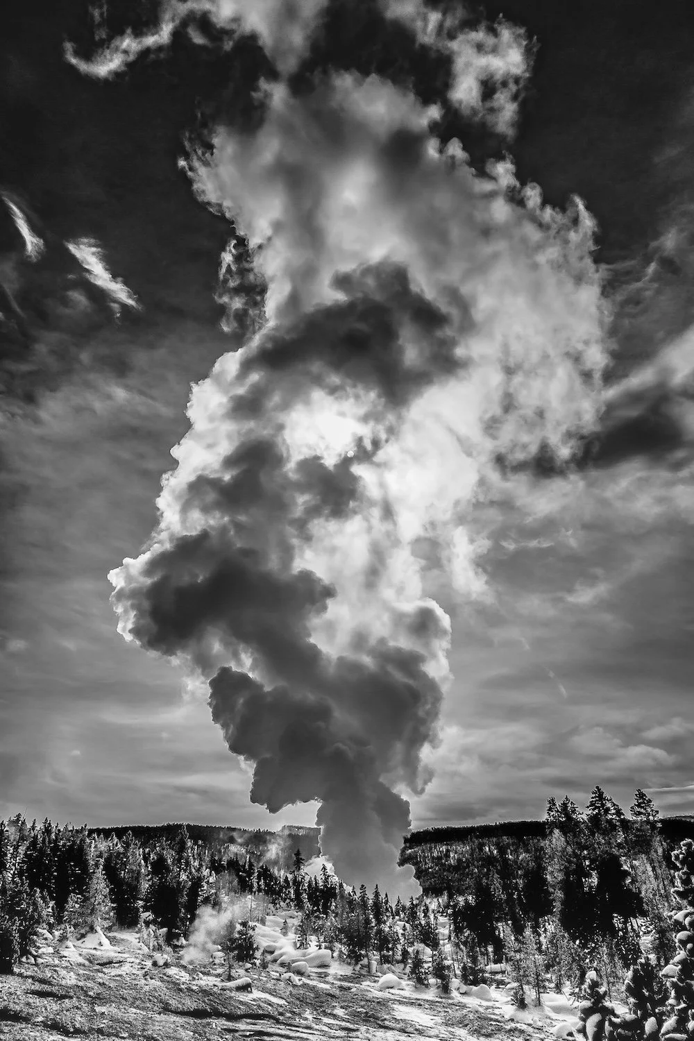 A black and white photograph of a Steamboat Geyser in Yellowstone National Park creates a large plume of steam rising into the sky, surrounded by a snow-covered forest landscape.