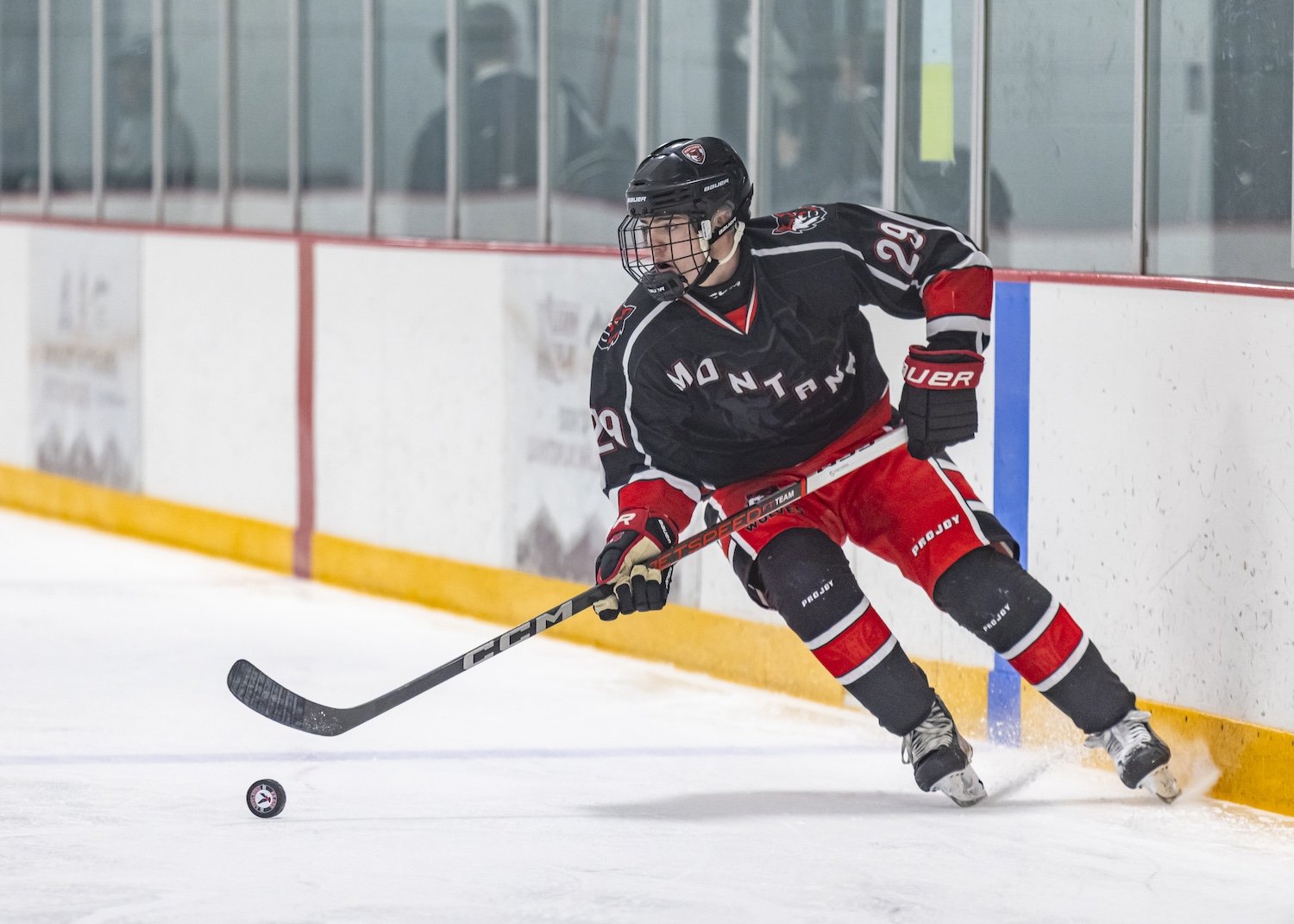 Ice hockey player in black and red uniform on ice rink, handling the puck near boards, wearing helmet and gloves.